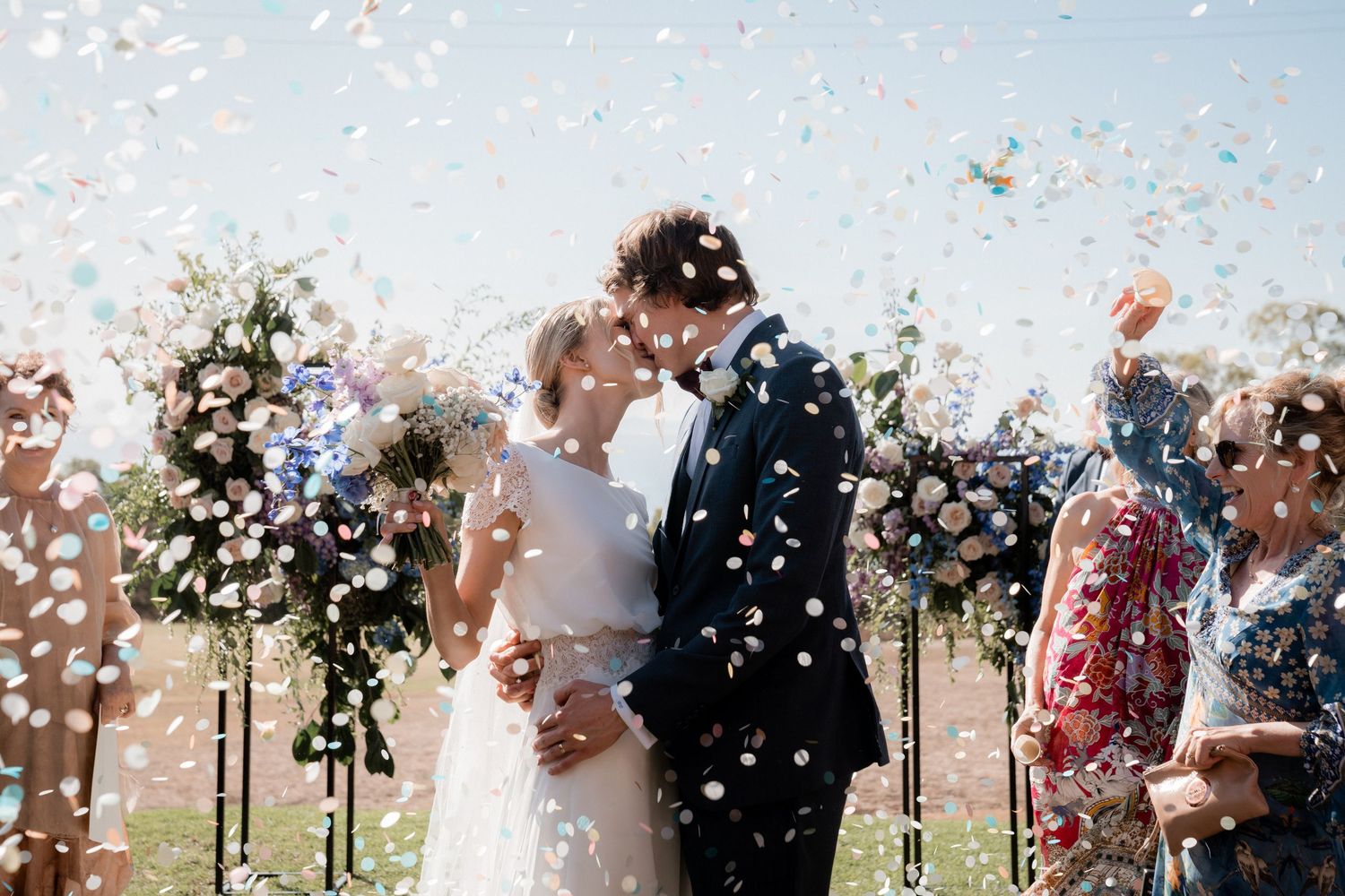 White confetti flutters around a couple during their outdoor wedding ceremony beneath floral arches.