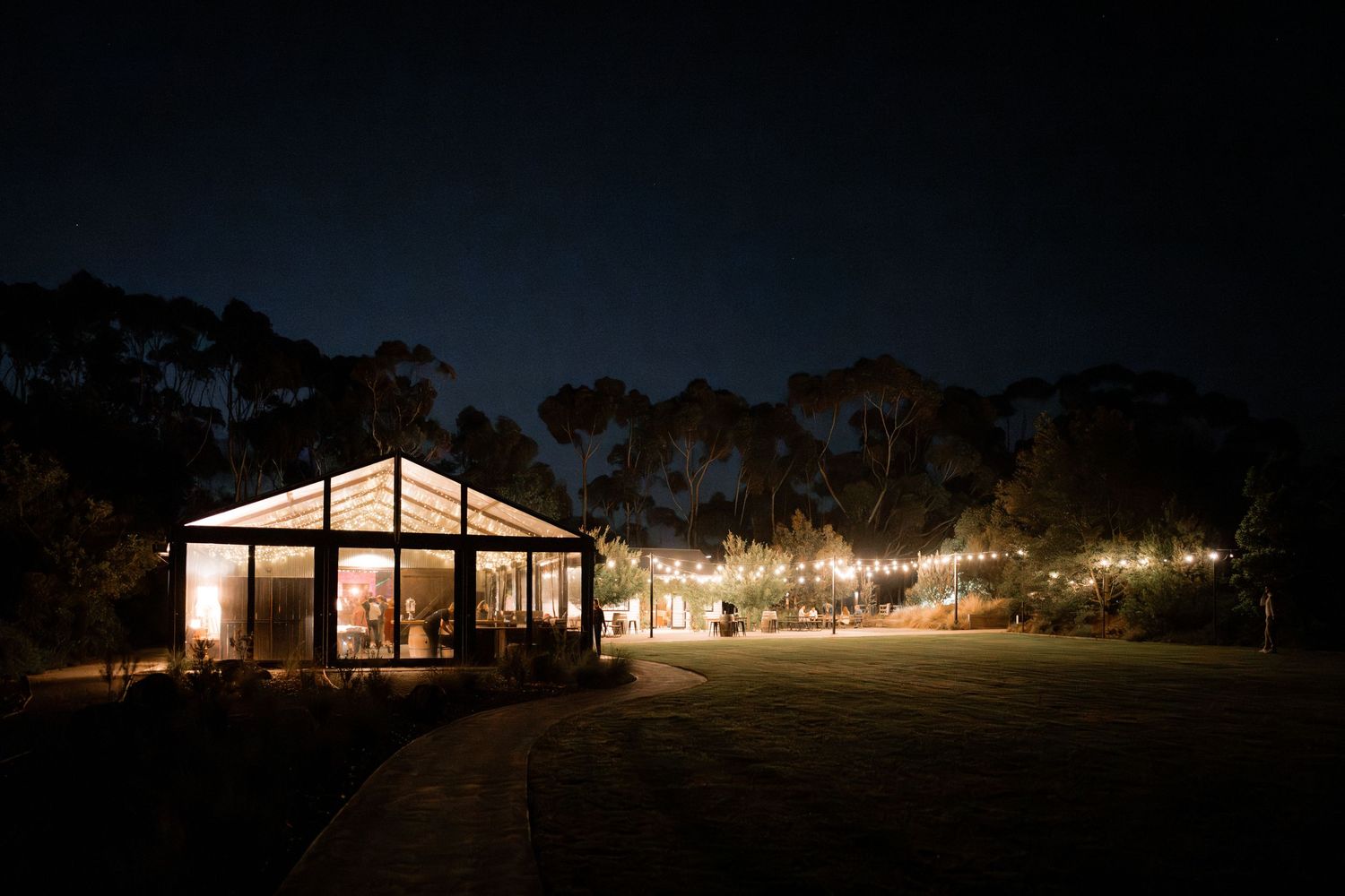 A clear-top tent illuminated at night with string lights leading to it across a dark outdoor venue.
