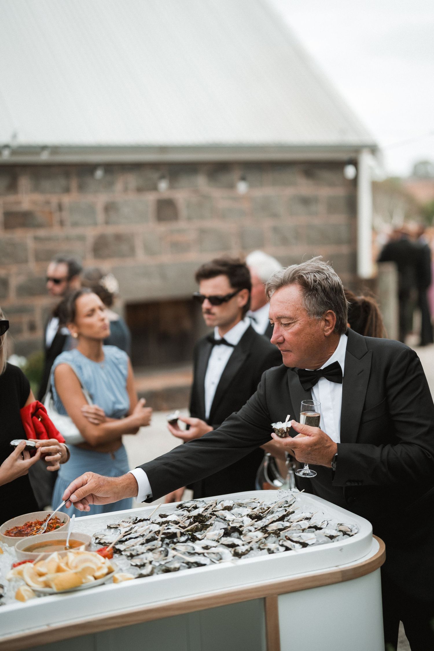 Guests in formal wear sample fresh oysters at an elegant outdoor wedding reception buffet.
