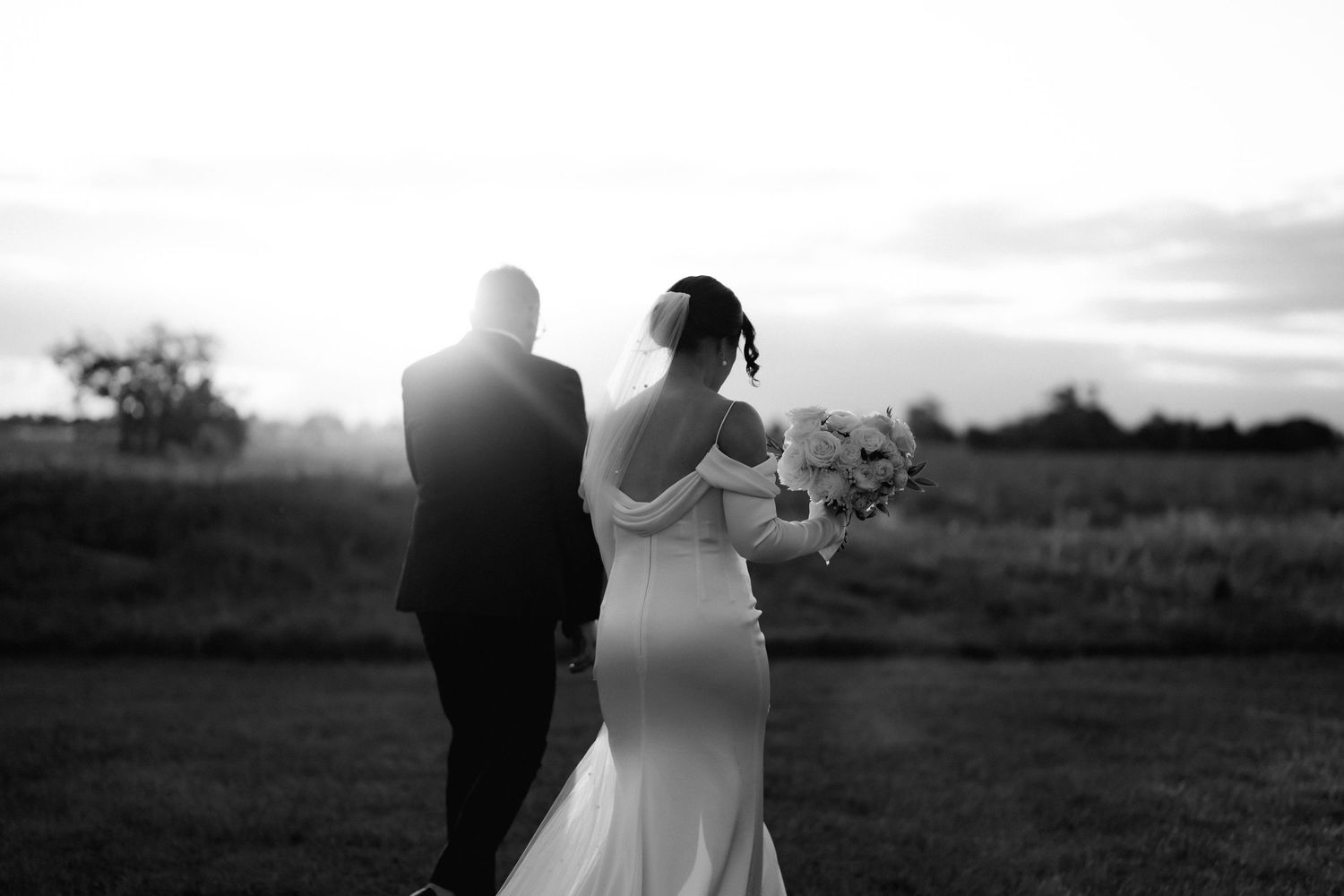 Newlyweds walk together at sunset in a field holding a bouquet.