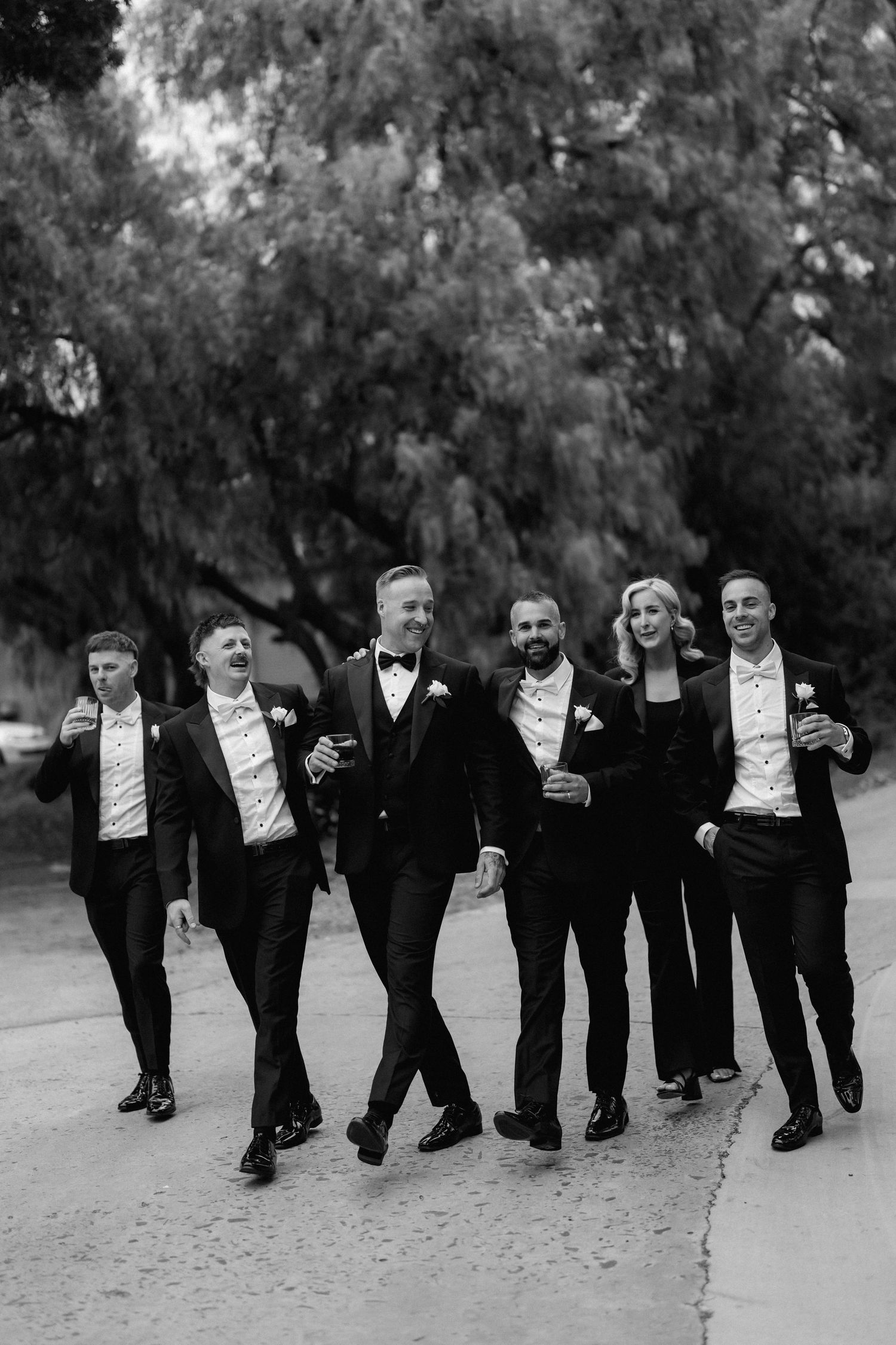 Group of groomsmen in tuxedos walking and laughing together on outdoor path in black and white photo.