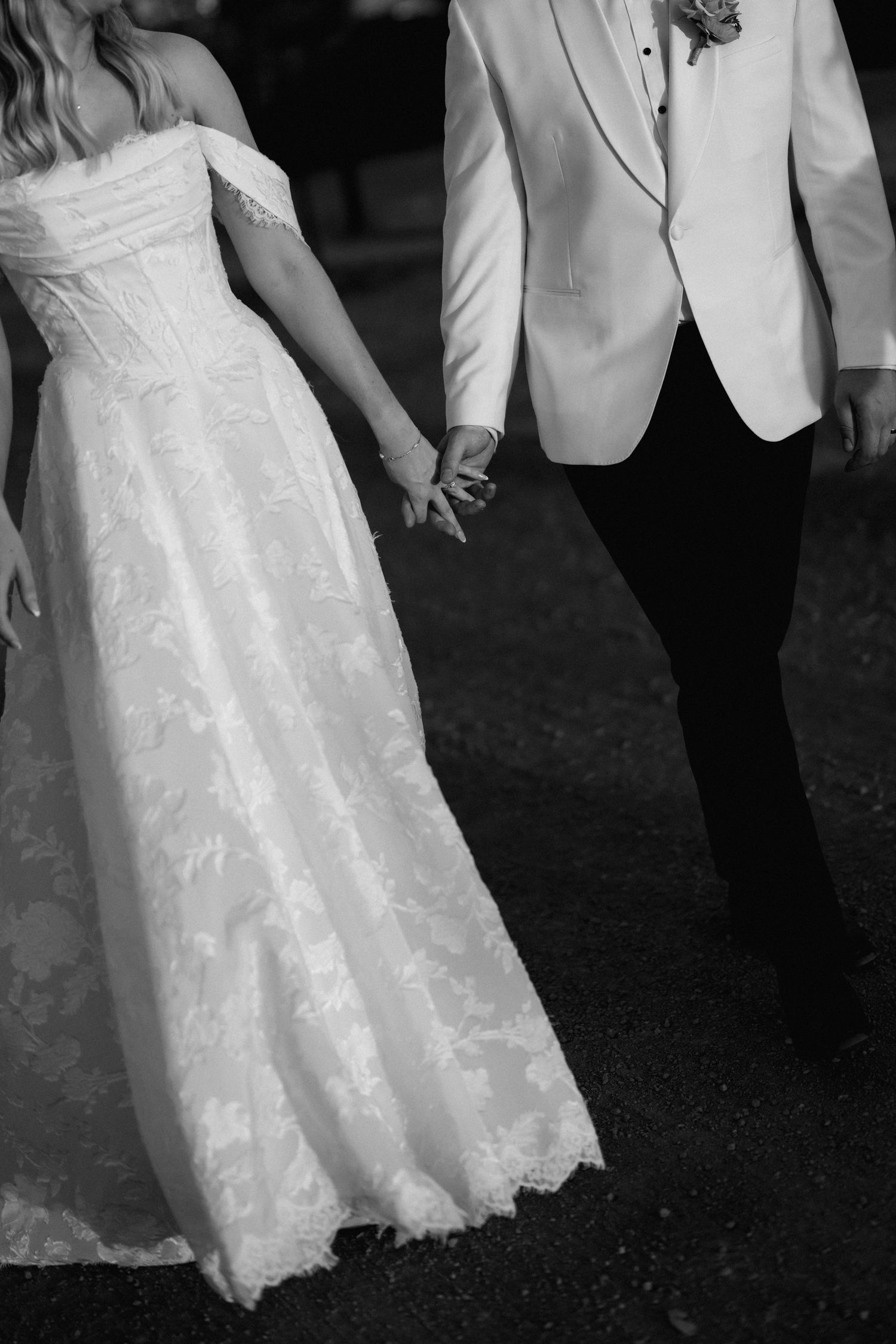 Black and white photo of bride in off-shoulder lace gown holding grooms hand while walking.