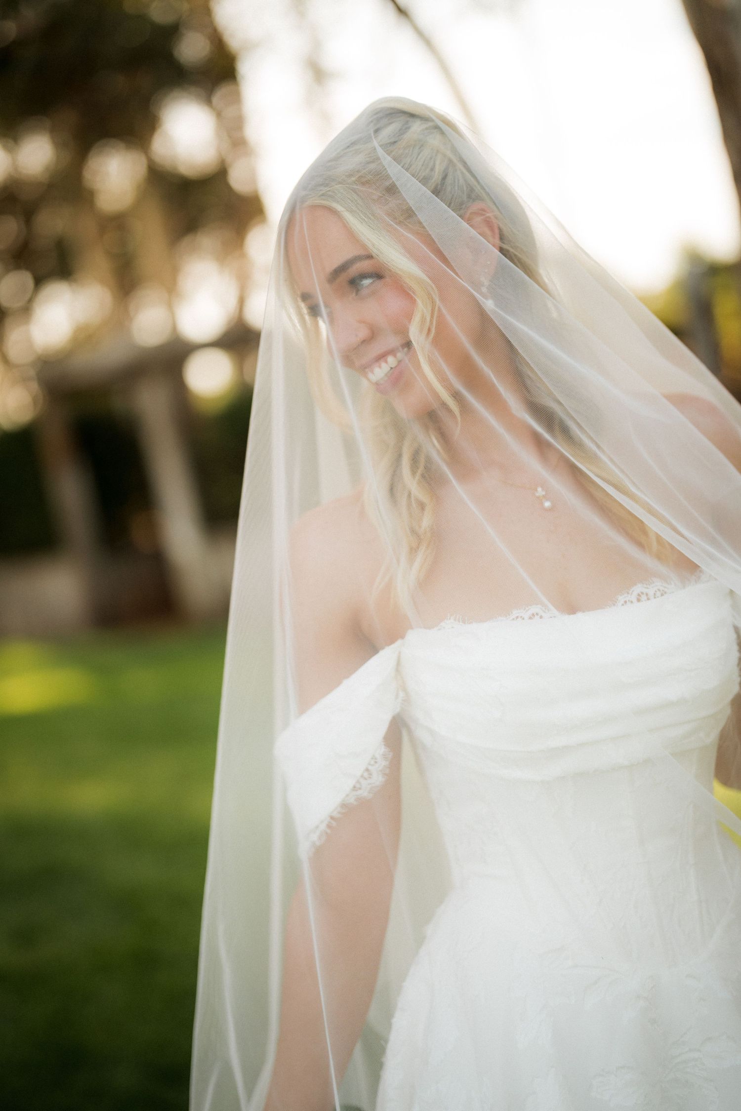 Smiling blonde bride in strapless lace gown draped in flowing veil outdoors.