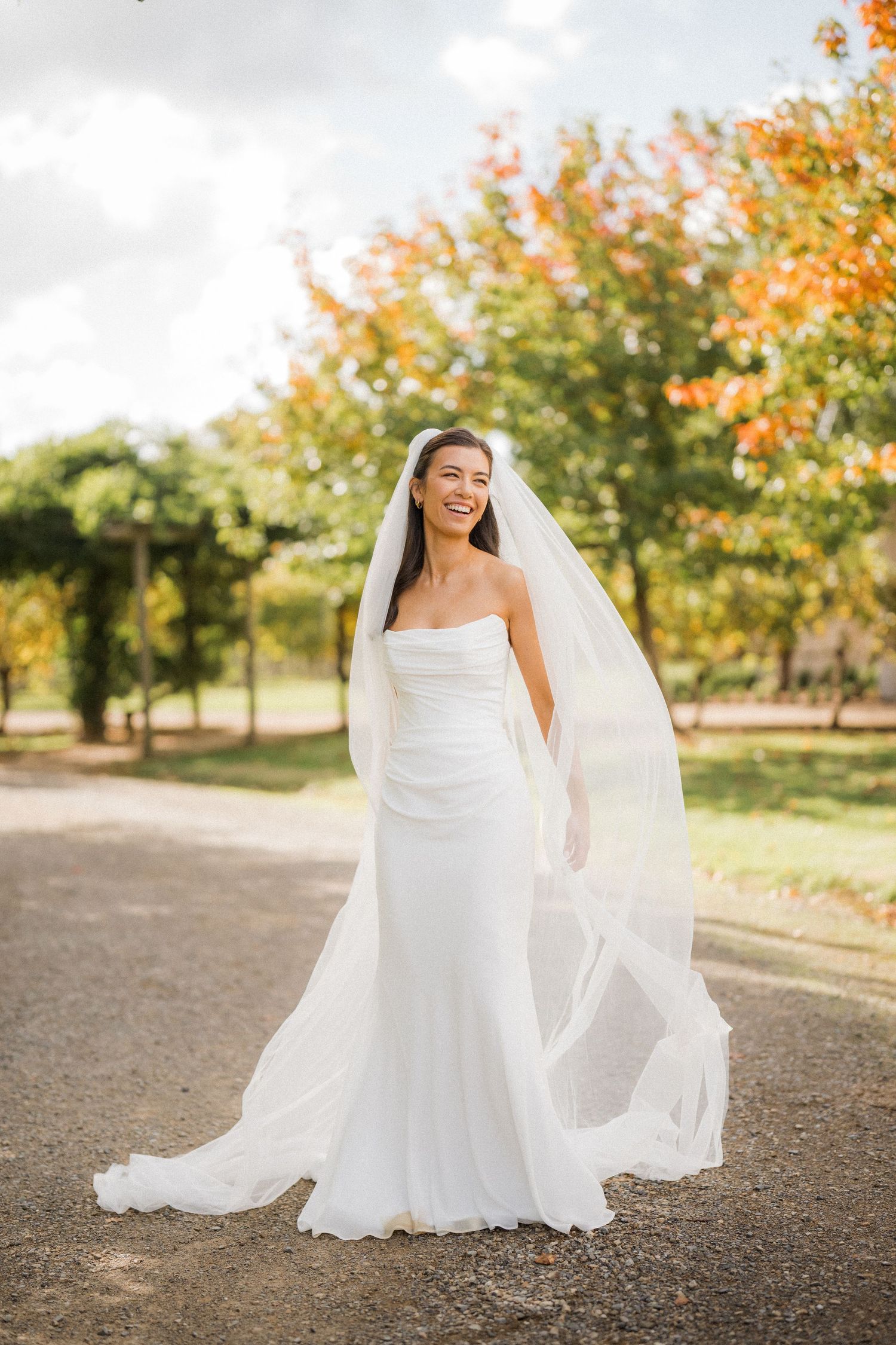 Radiant bride in strapless white gown with flowing veil standing on garden path in autumn.