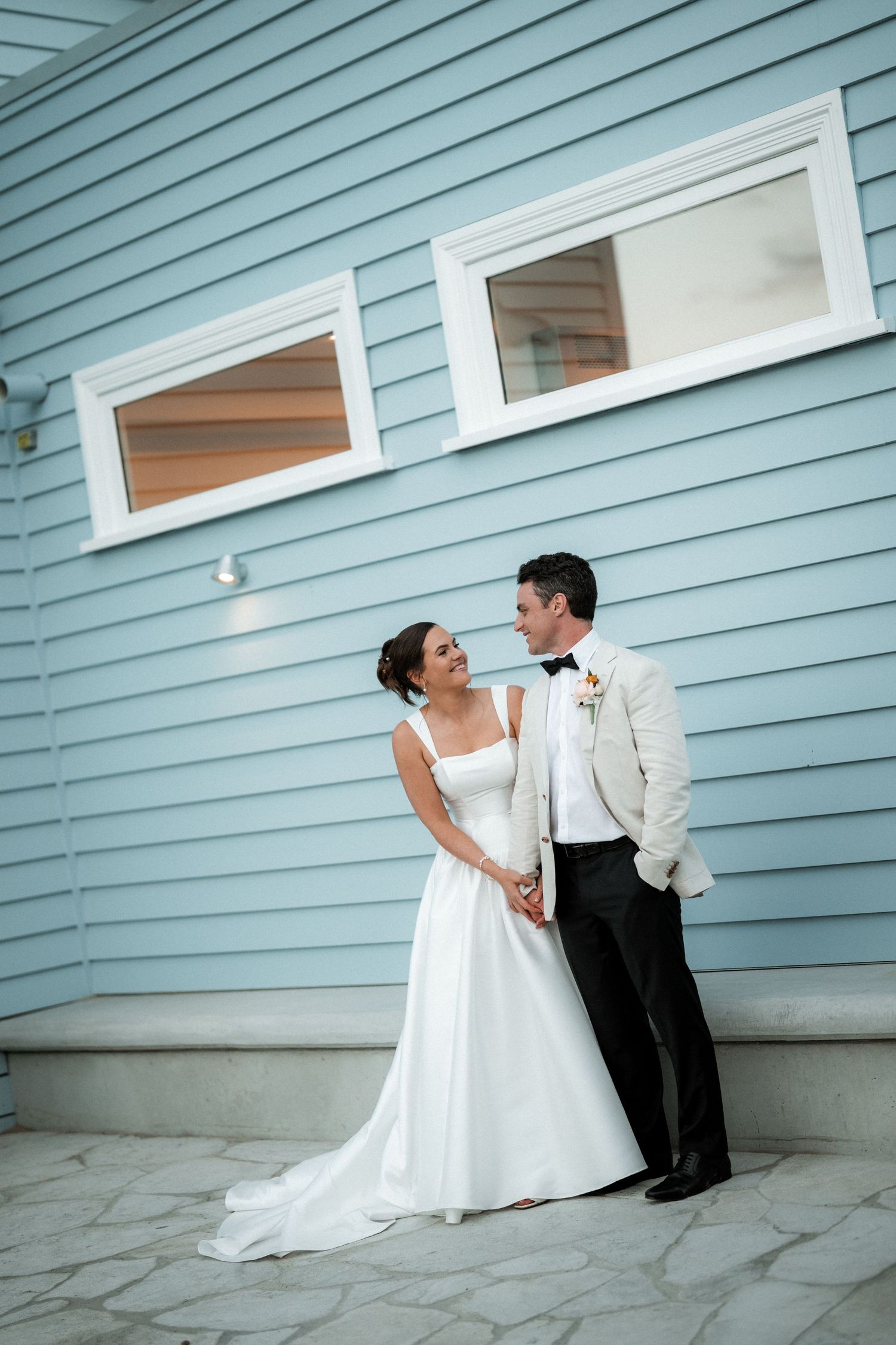 Newlyweds pose against a light blue siding of a coastal home in elegant wedding attire.