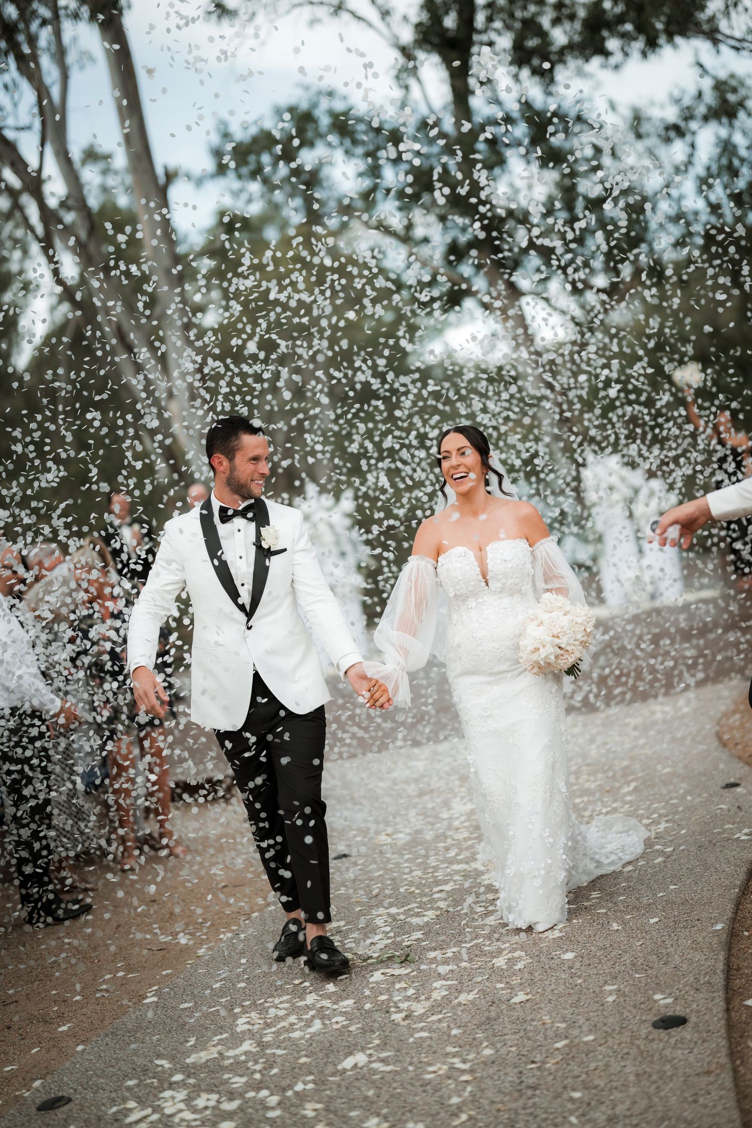 Wedding couple running through a shower of white confetti on a garden path.