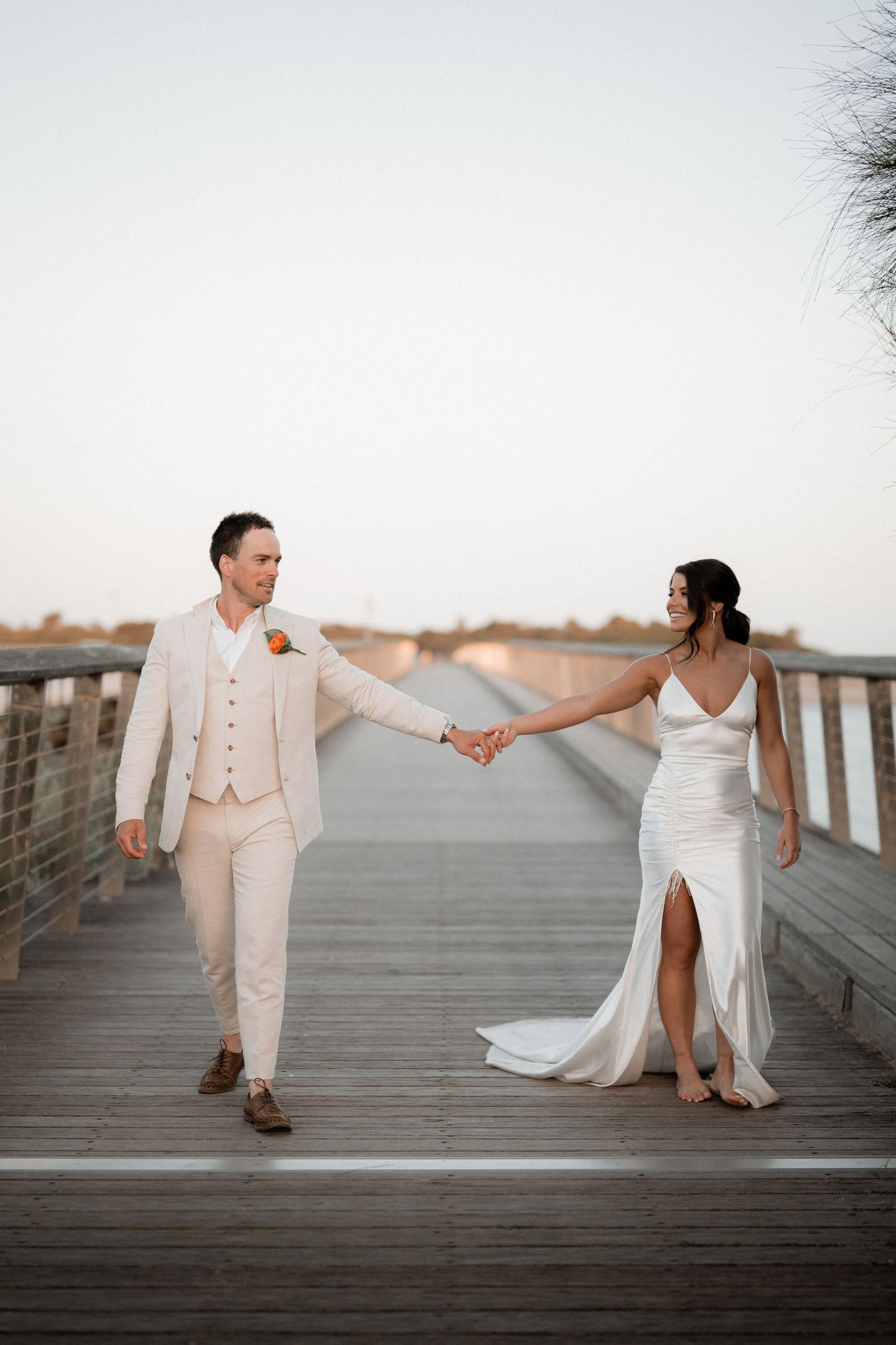 Couple in wedding attire walking hand in hand on wooden boardwalk at sunset.