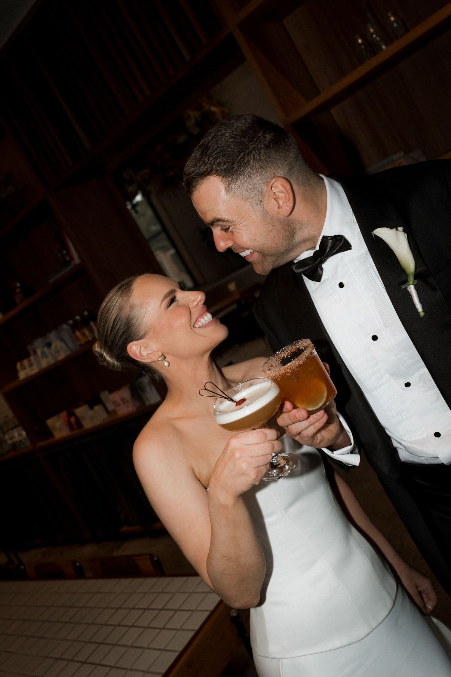 Newlyweds share drinks and laughter during wedding reception in intimate bar setting.