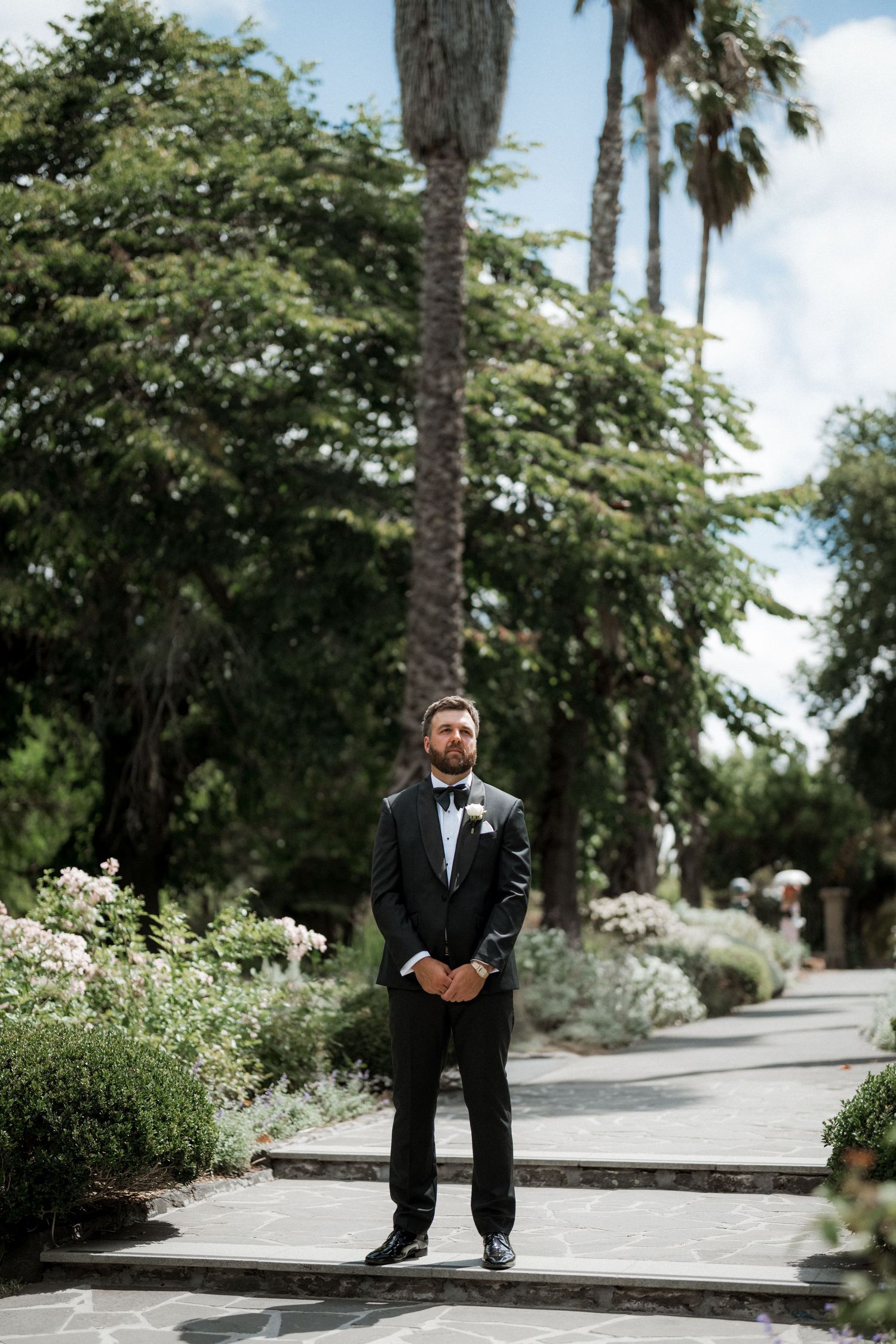 A dapper groom in a black tuxedo with bow tie stands confidently on a stone path surrounded by lush garden greenery.