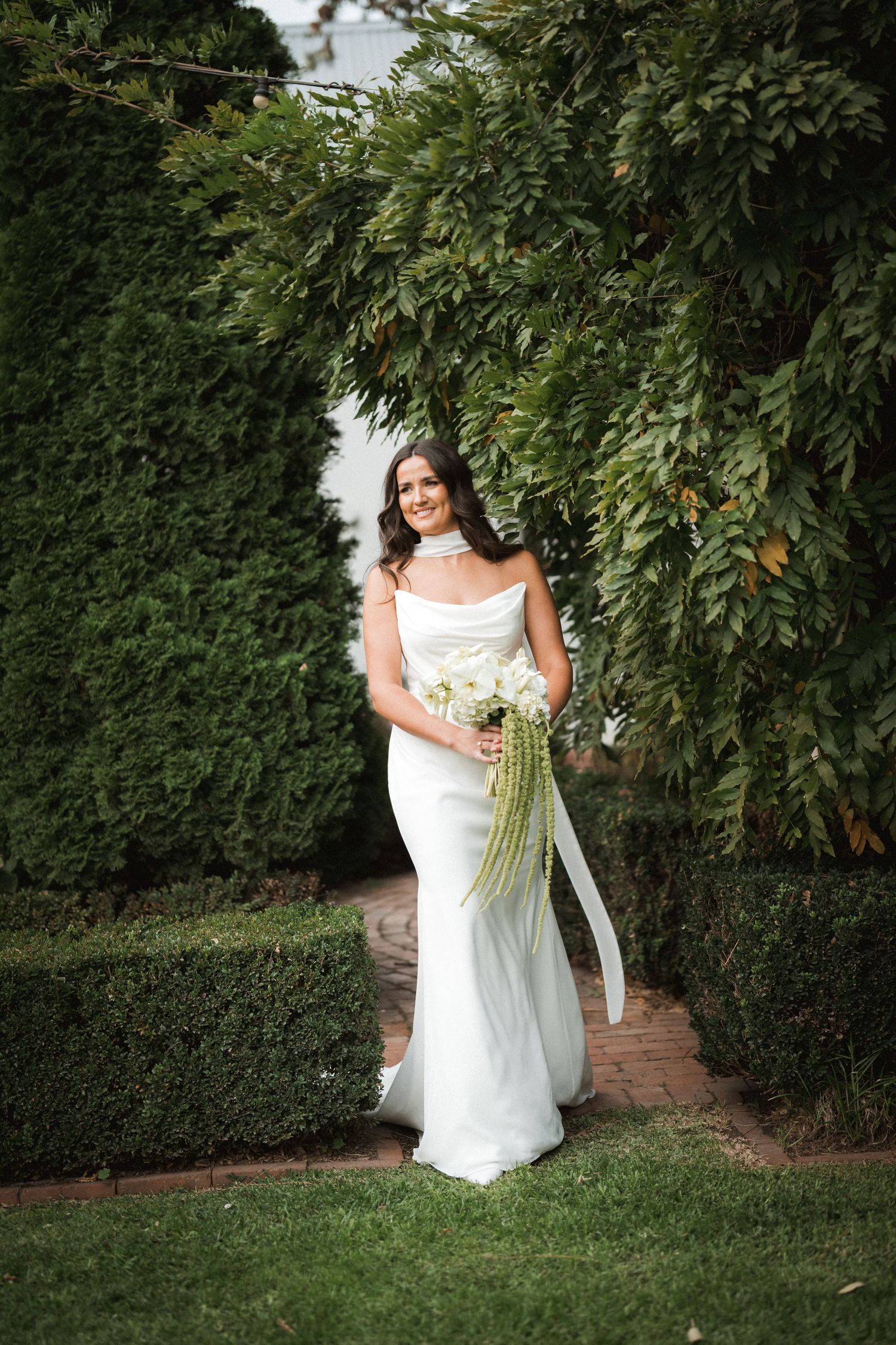 Bride in white dress holding bouquet poses against lush garden hedge and brick wall.