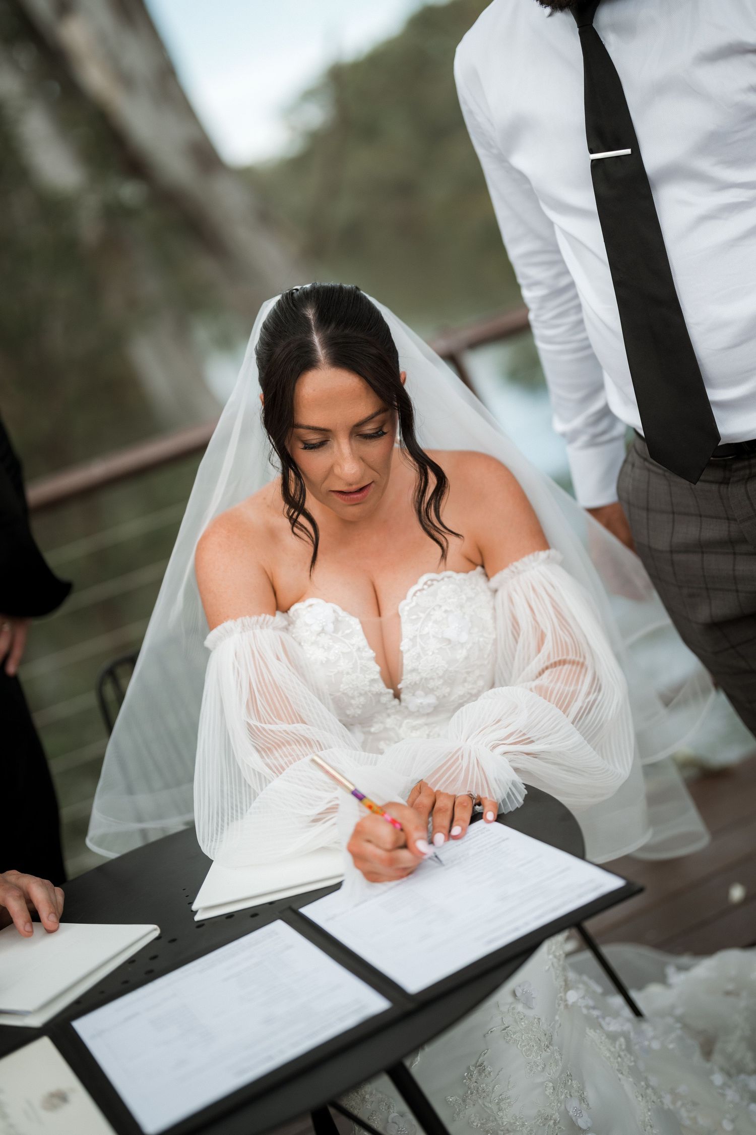 Bride in white lace dress signs marriage documents during outdoor wedding ceremony.
