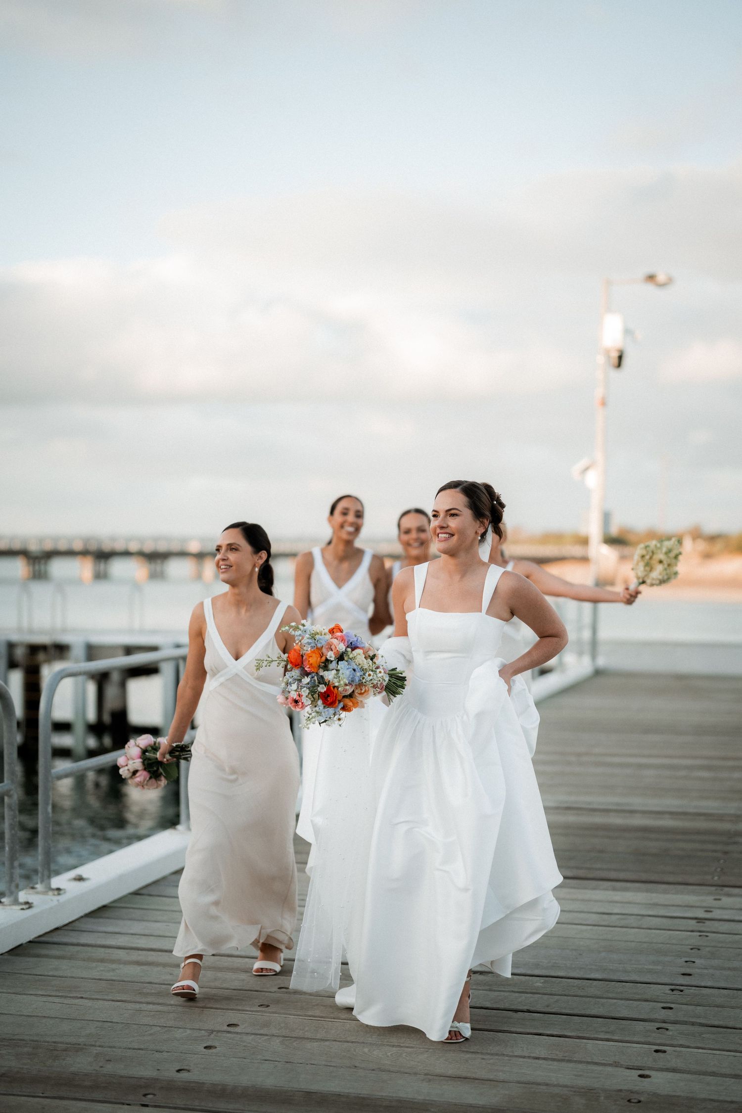 Wedding party members dance and celebrate on a wooden boardwalk by the ocean.