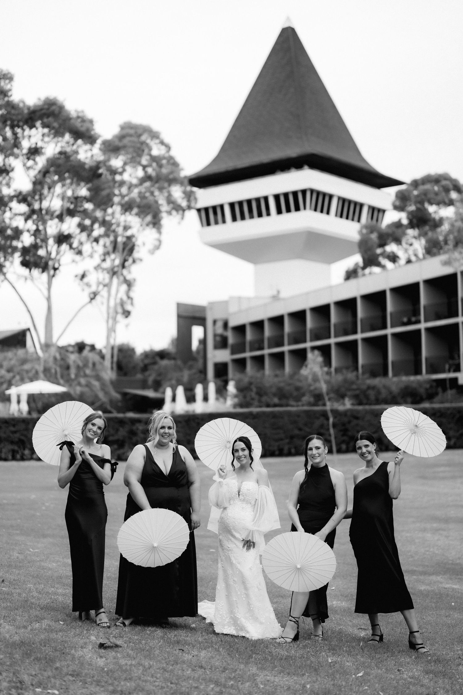Wedding party with parasols posing on lawn in front of a building with distinctive tower architecture.