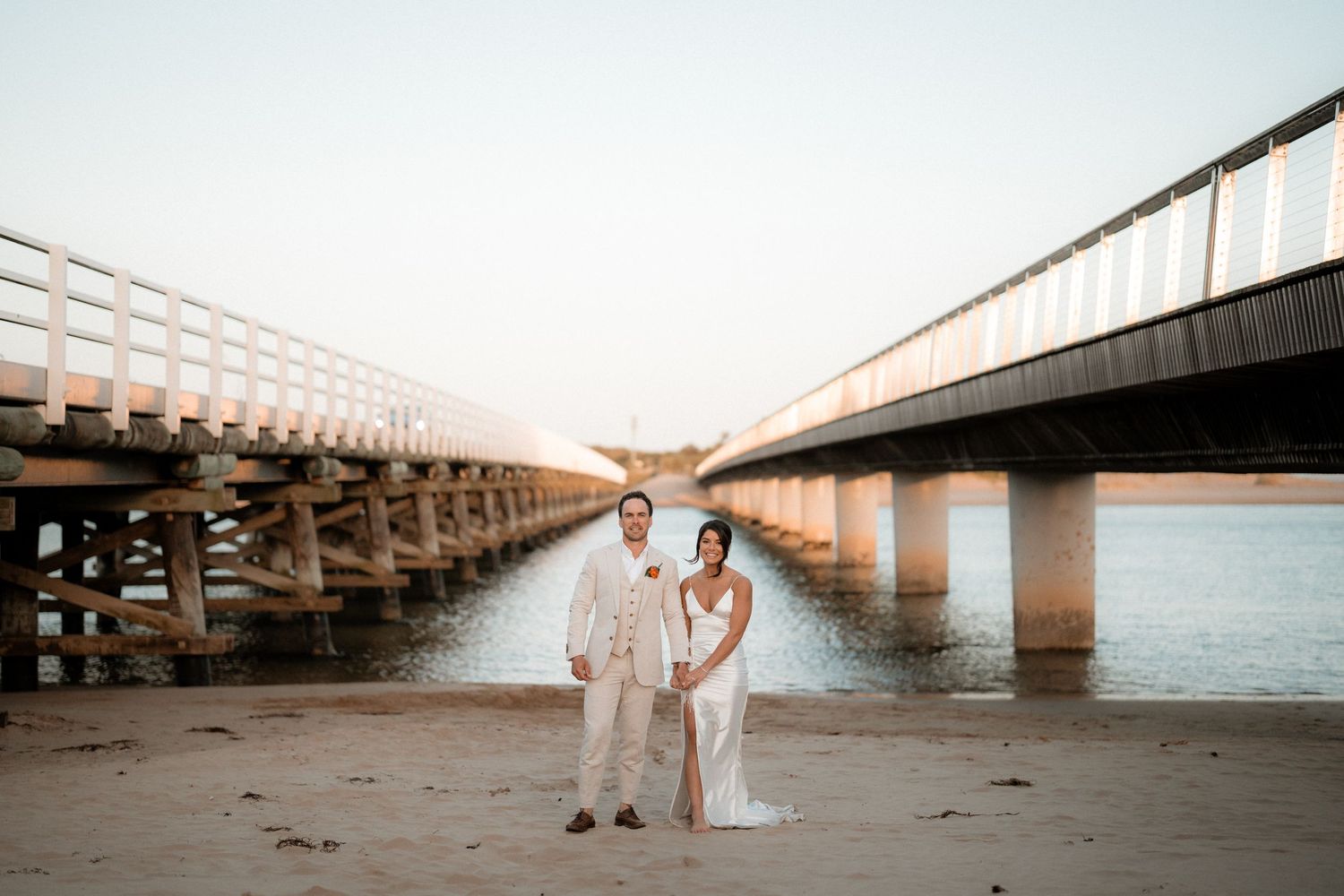 Newlyweds pose for romantic photos beneath a long bridge on a sandy beach at sunset.