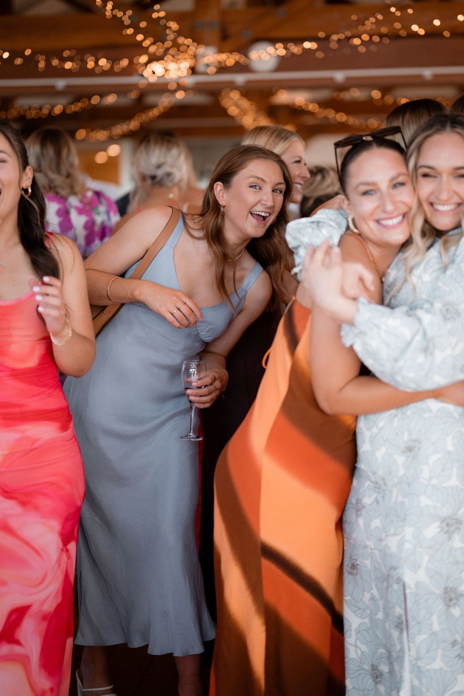 Wedding party dancing and celebrating in colorful dresses under indoor lighting.