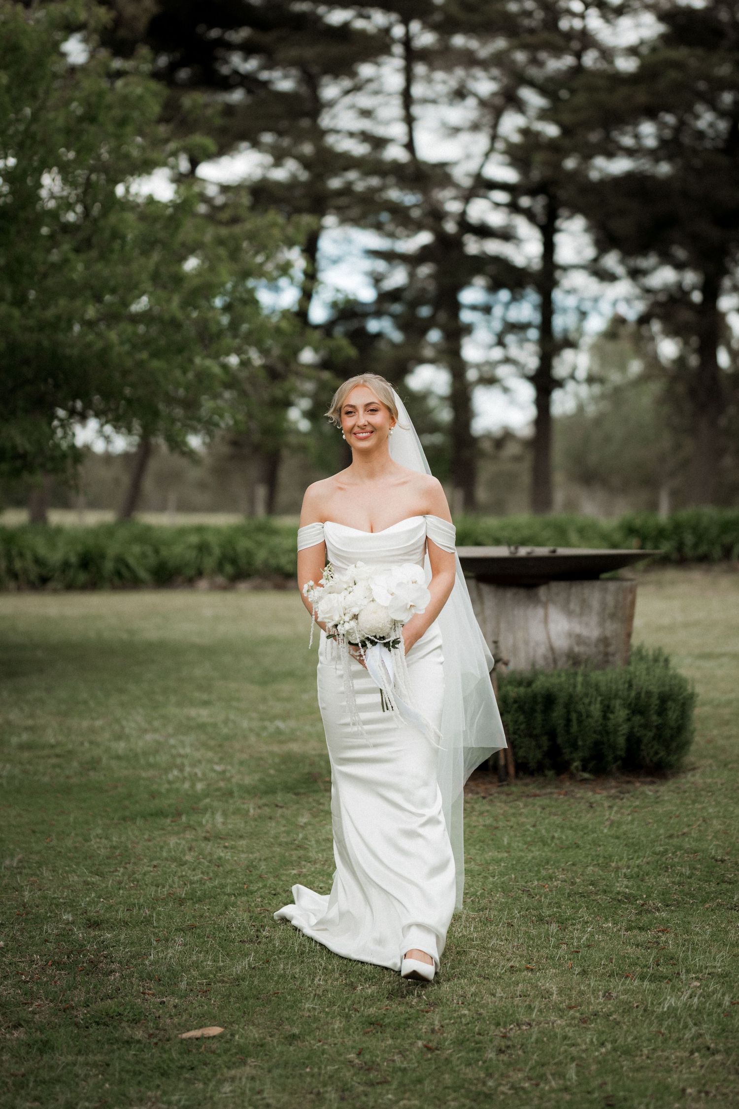 Joyful bride in white dress walking and smiling on grassy lawn with trees in background.