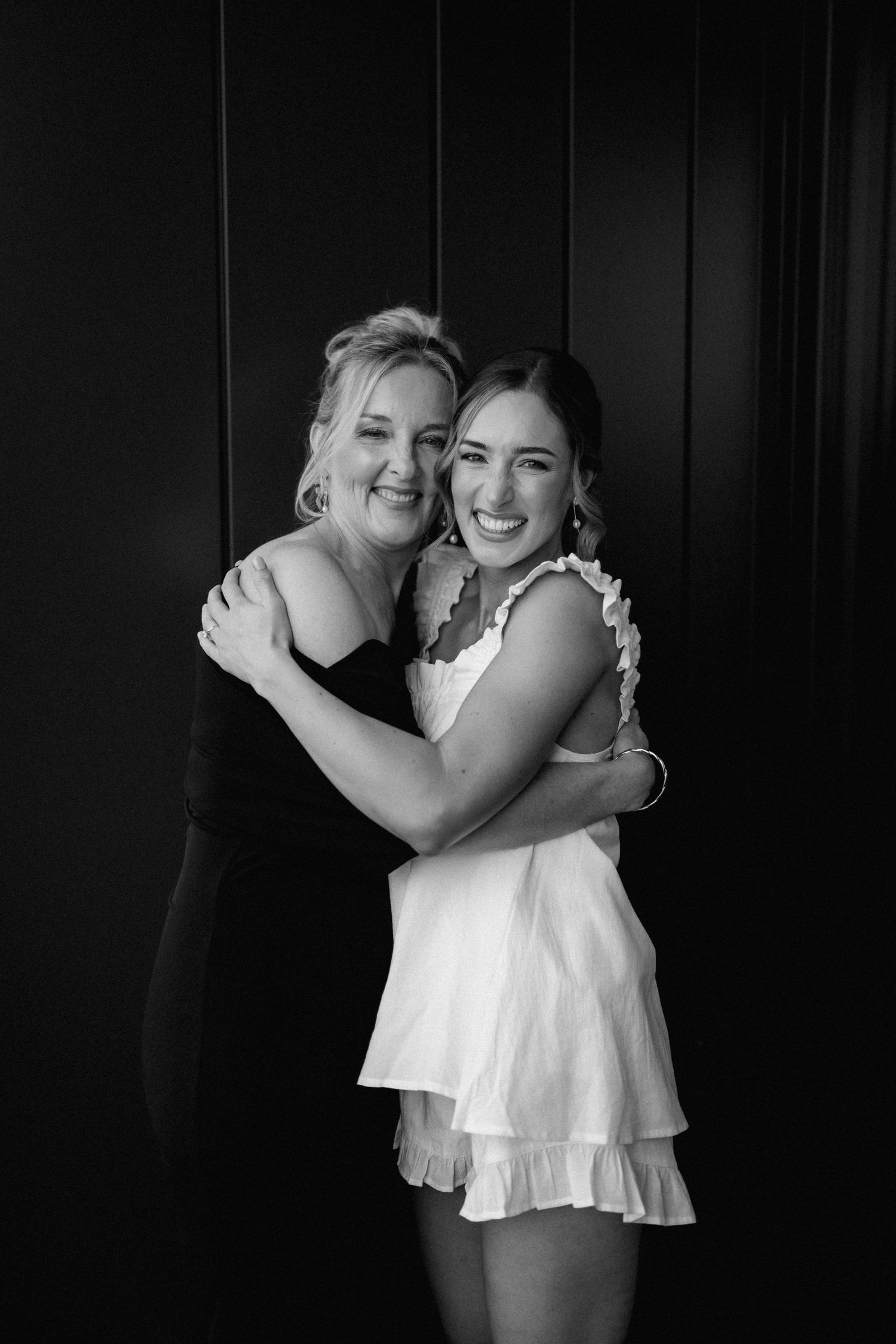 Black and white portrait of two women in contrasting black and white outfits embracing and smiling against dark background.
