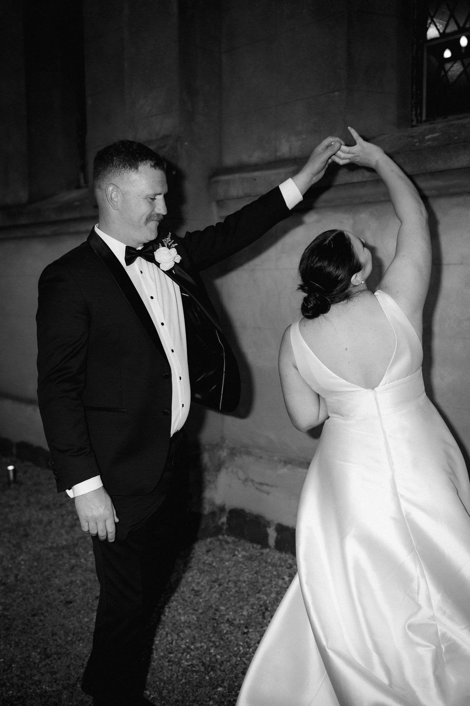 A couple shares an elegant dance moment in formal attire, with the bride in a white gown and groom in a black tuxedo.
