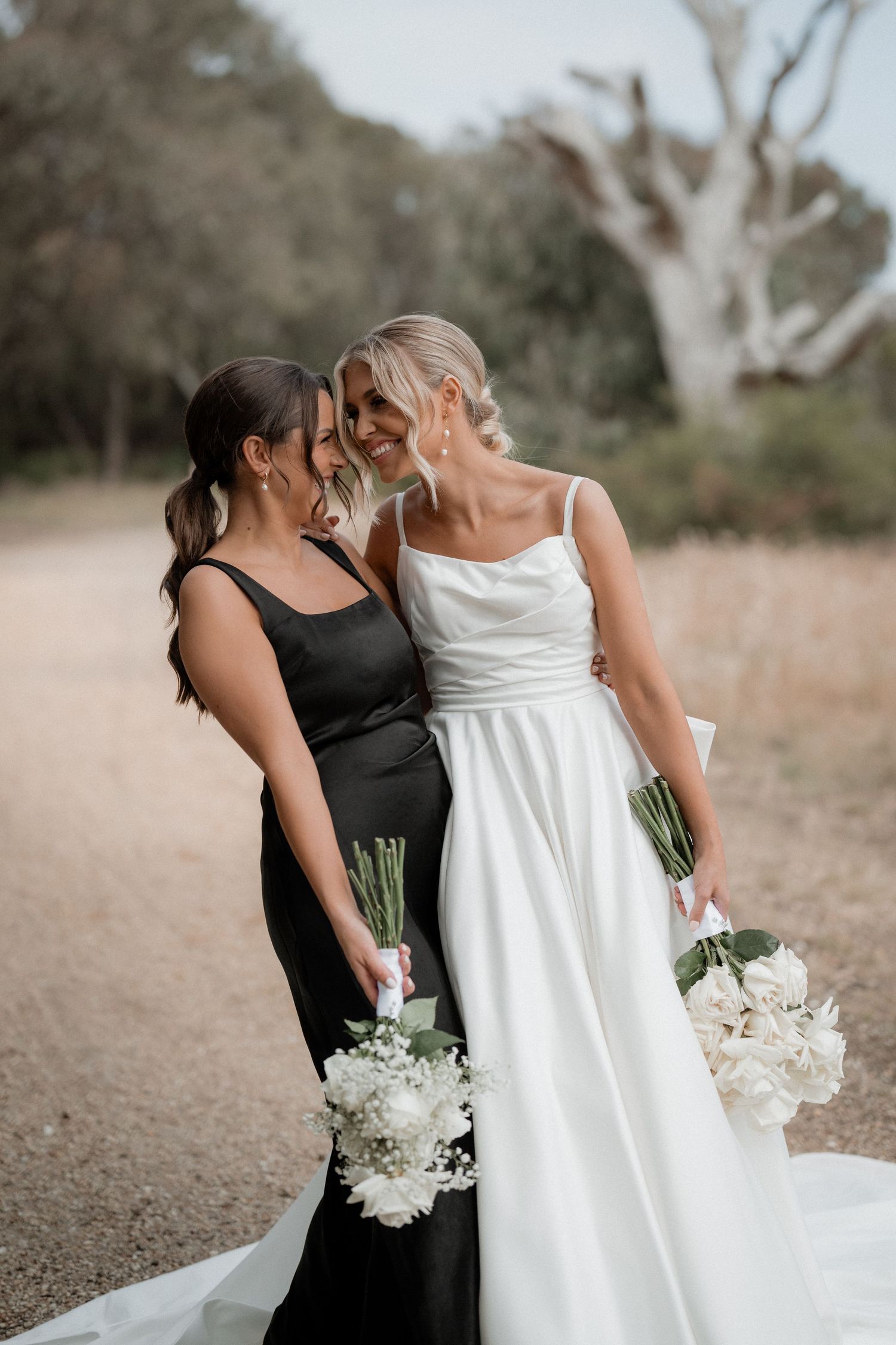 Two people in contrasting black and white dresses share an intimate moment while holding white bouquets on a dirt path.