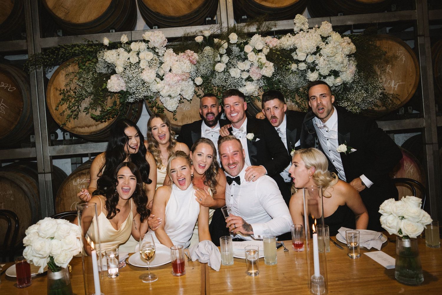 A wedding party gathers at a rustic venue with wine barrels and white hydrangea floral arrangements.