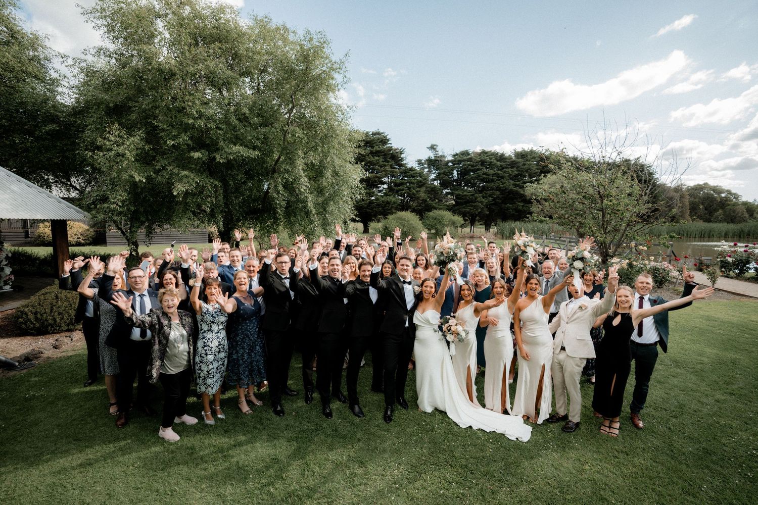 Large wedding party gathered on a lush green lawn with trees in the background during an outdoor celebration.