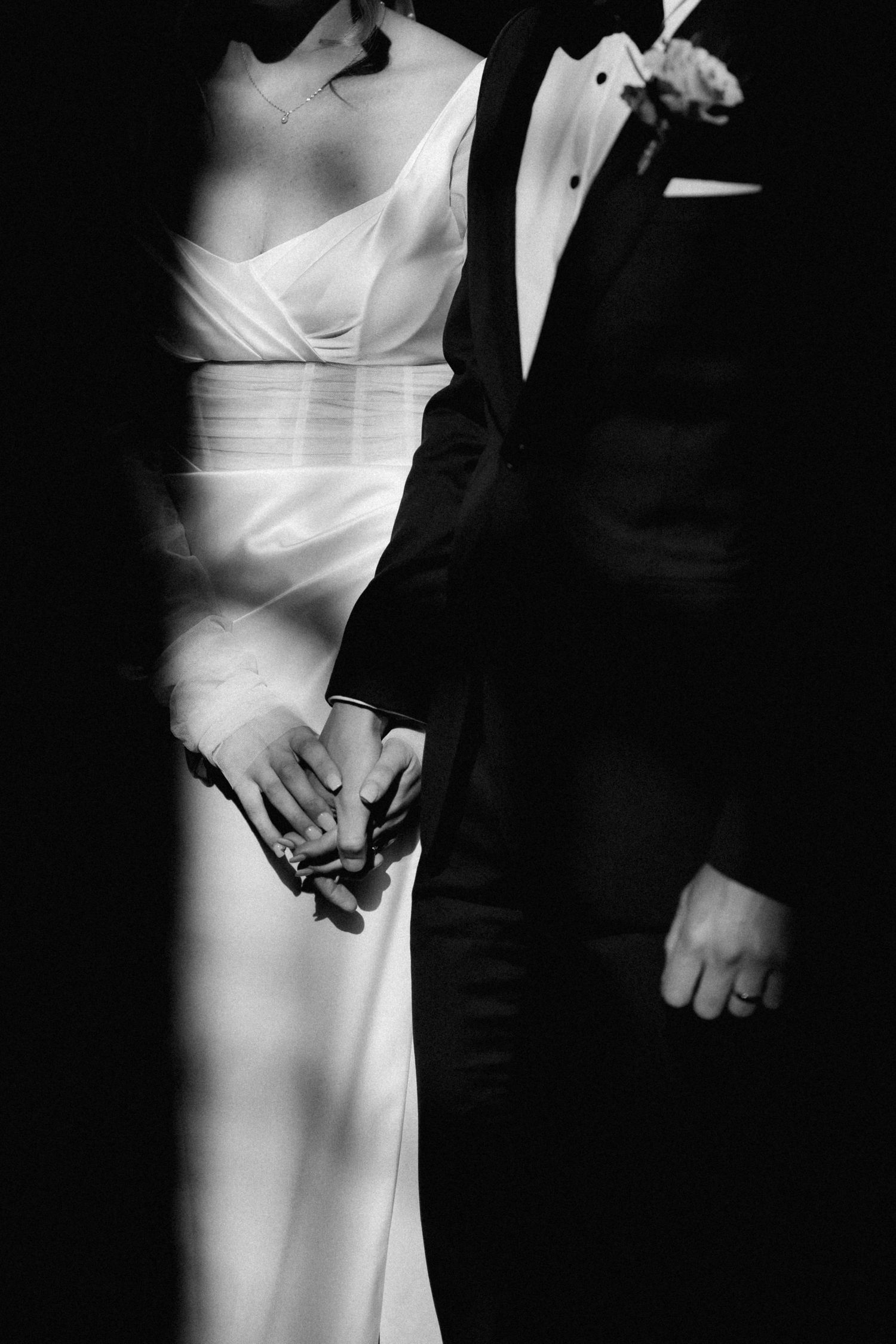 Black and white photo of couple in formal wedding attire holding hands in dramatic lighting.