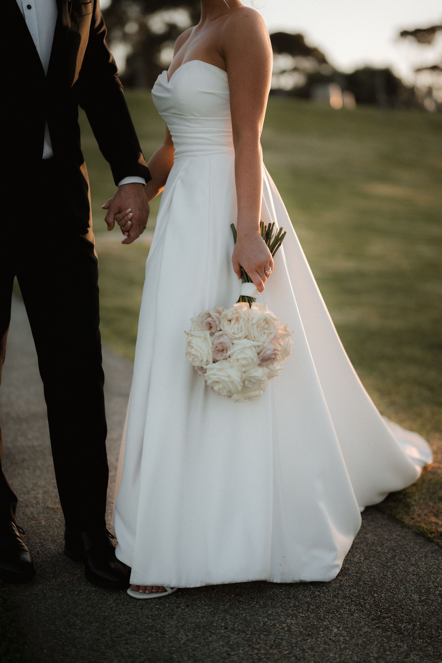 Detail shot of couple holding hands with bride's flowing white dress and bouquet against garden backdrop.