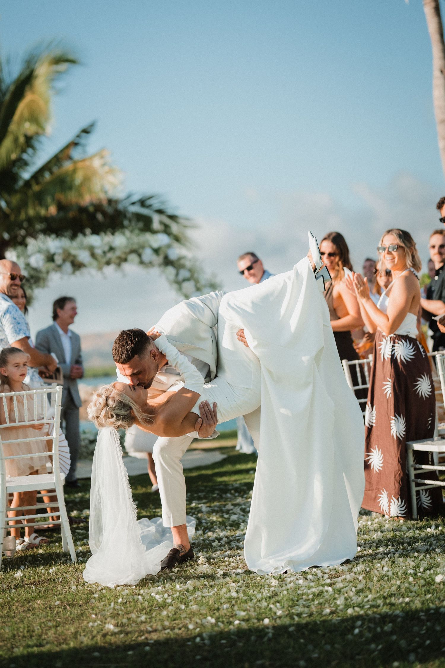 A joyful outdoor wedding celebration with people gathered on a sunny tropical beach with palm trees in the background.