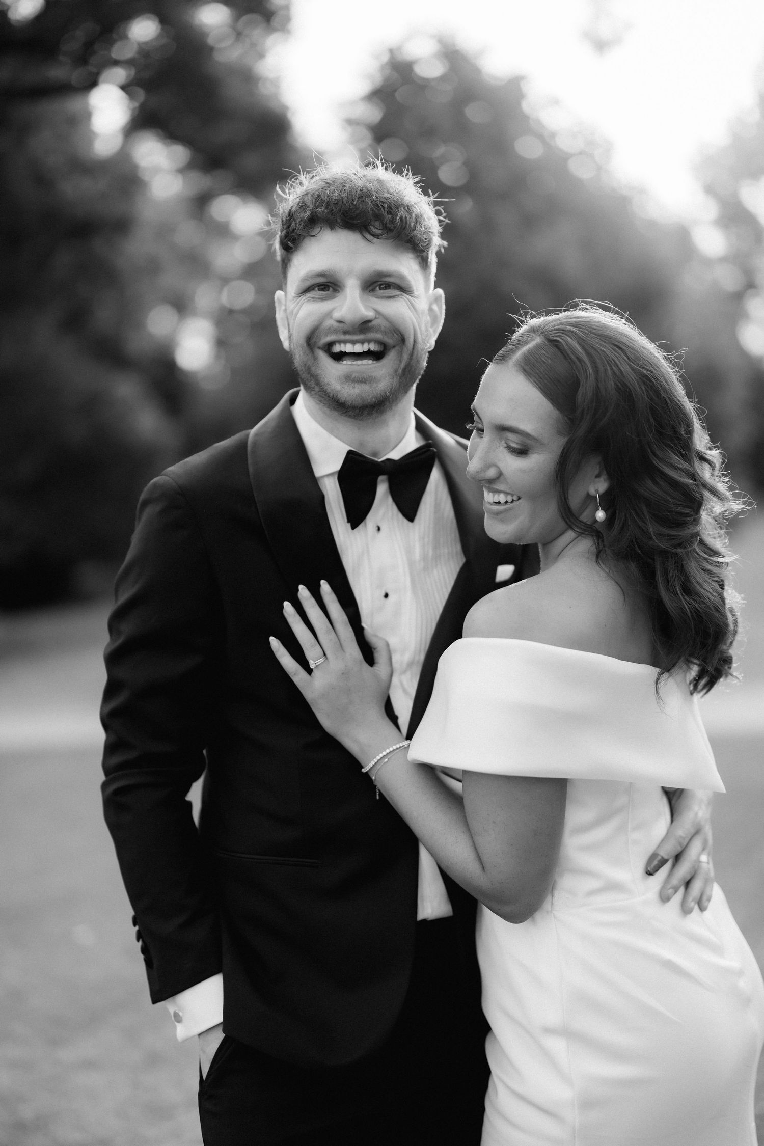 Candid black and white portrait of a newlywed couple in formal attire sharing a joyful moment outdoors.