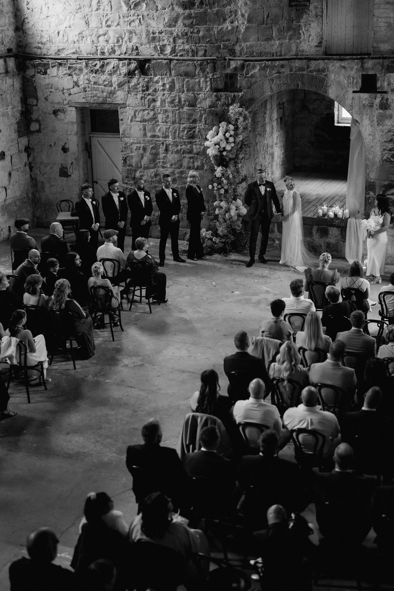 Black and white photo of wedding ceremony in rustic stone venue with guests seated in rows.
