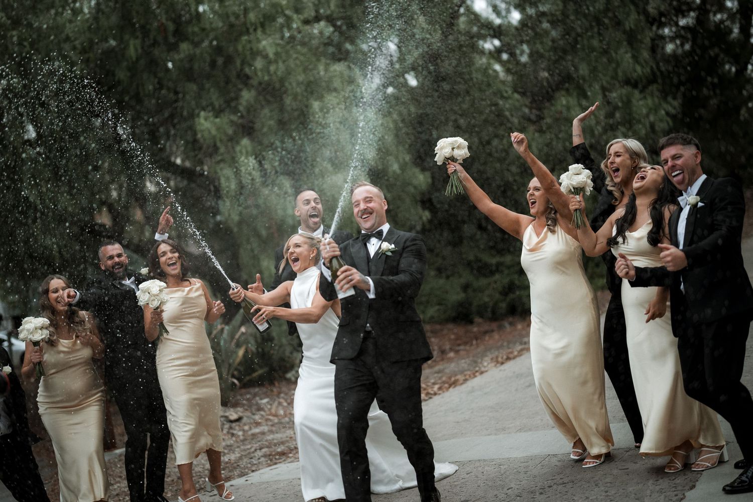 Wedding party celebrating outdoors with champagne spraying and joyful expressions on a woodland path.