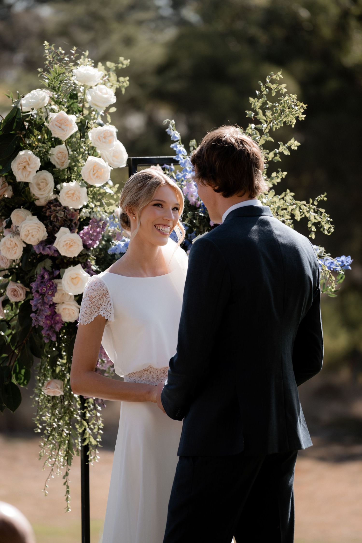 A beautiful outdoor ceremony features a couple standing at a floral arch with white roses and greenery.
