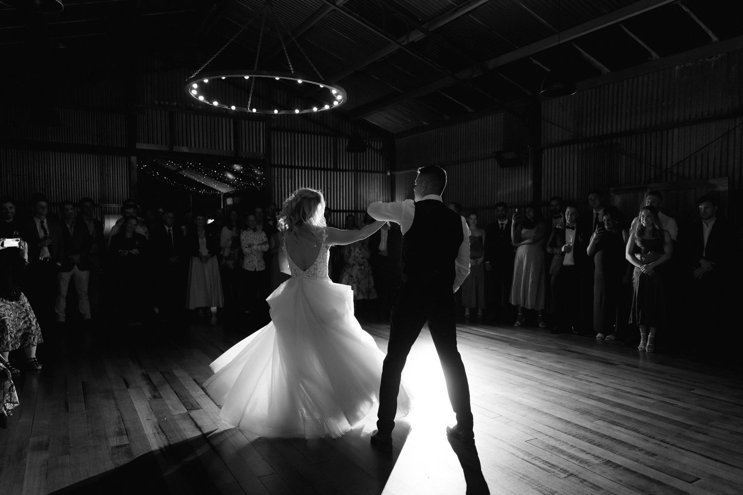 A couple performs their first dance as newlyweds in a dimly lit reception hall with twirling silhouettes on the dance floor.