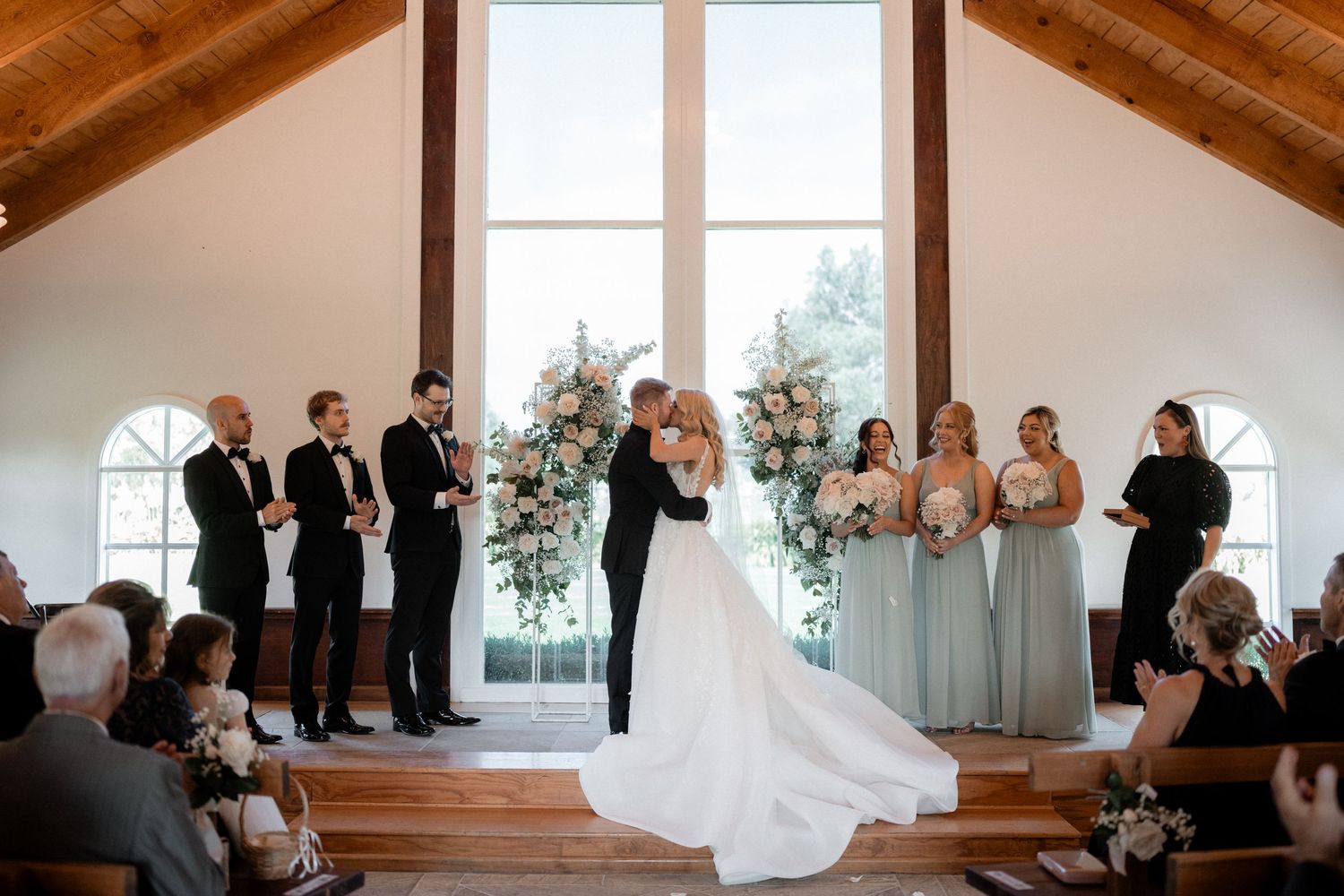 Wedding ceremony takes place in a bright chapel with wooden beams and arched windows as people gather to witness the occasion.