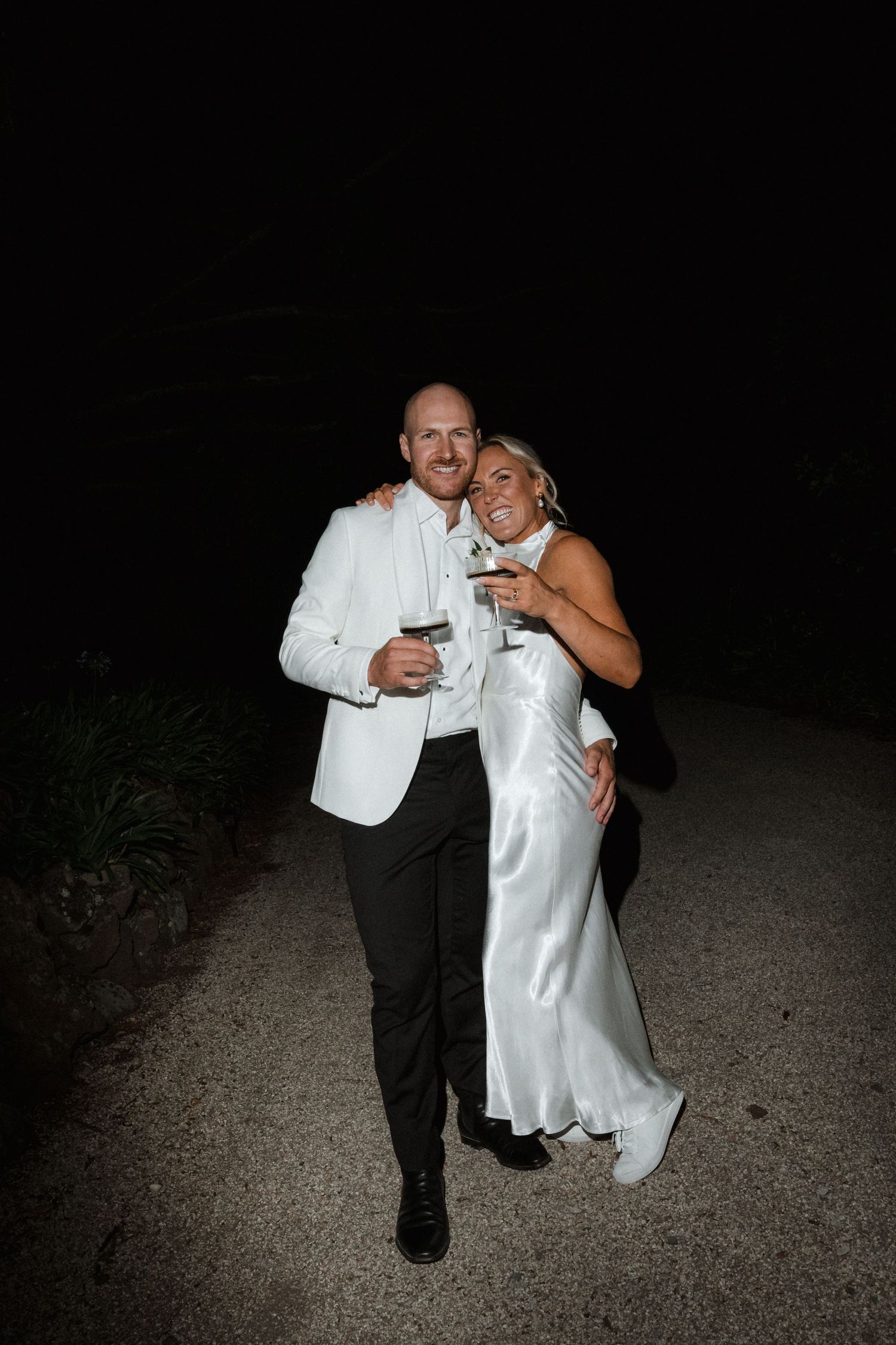 A romantic nighttime photo of newlyweds sharing drinks and laughing together on a dark pathway.