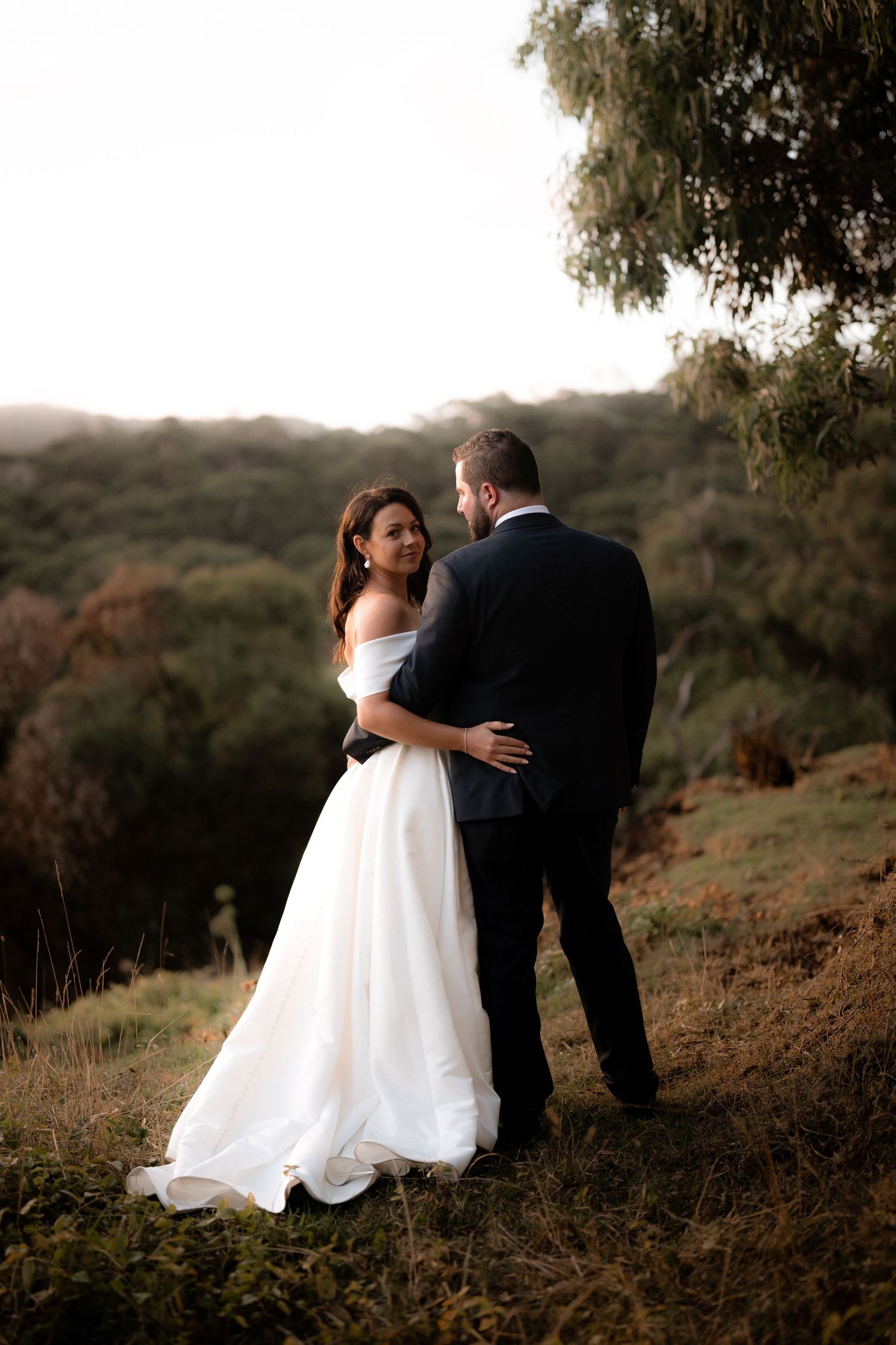 A romantic wedding couple embraces on a hilltop at sunset with a misty forest backdrop.