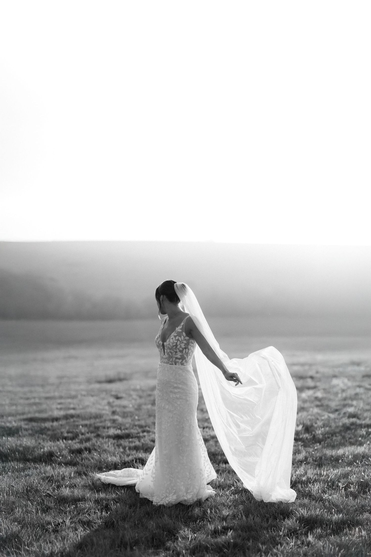 Black and white photo of a bride in a lace gown with flowing veil standing in an open field.