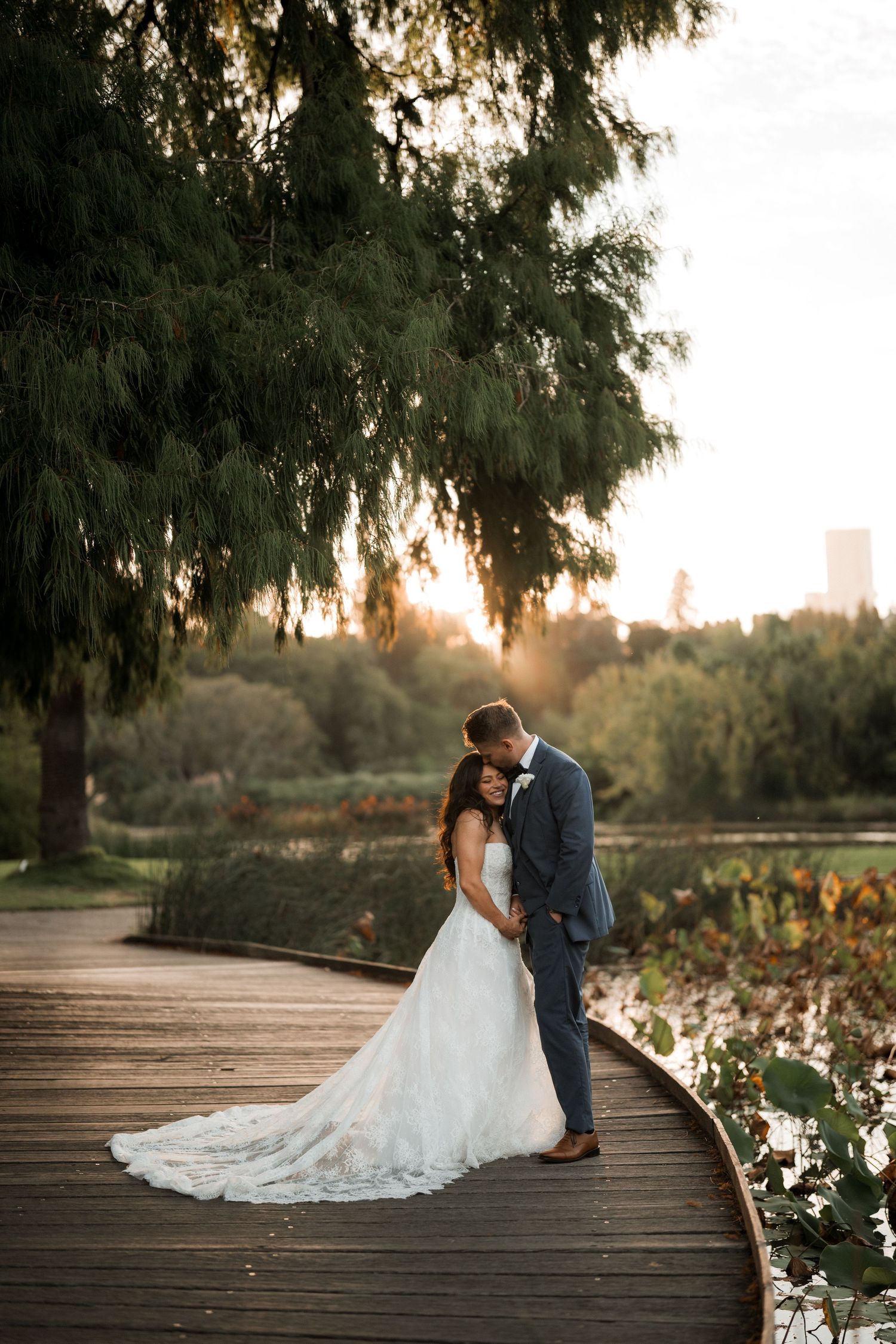 Romantic sunset silhouette of a couple in wedding attire sitting on a wooden dock by a tranquil lake with trees.