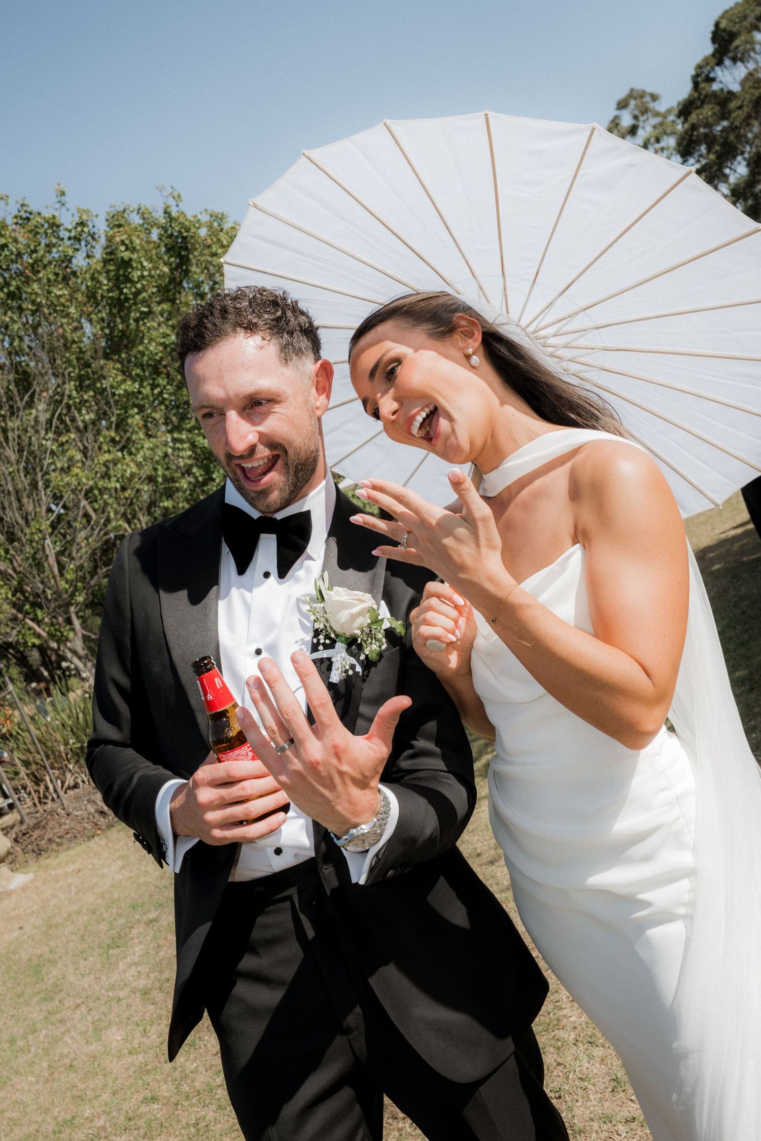 A joyful wedding moment outdoors with a white parasol, bouquet, tuxedo and elegant white gown on a sunny day.