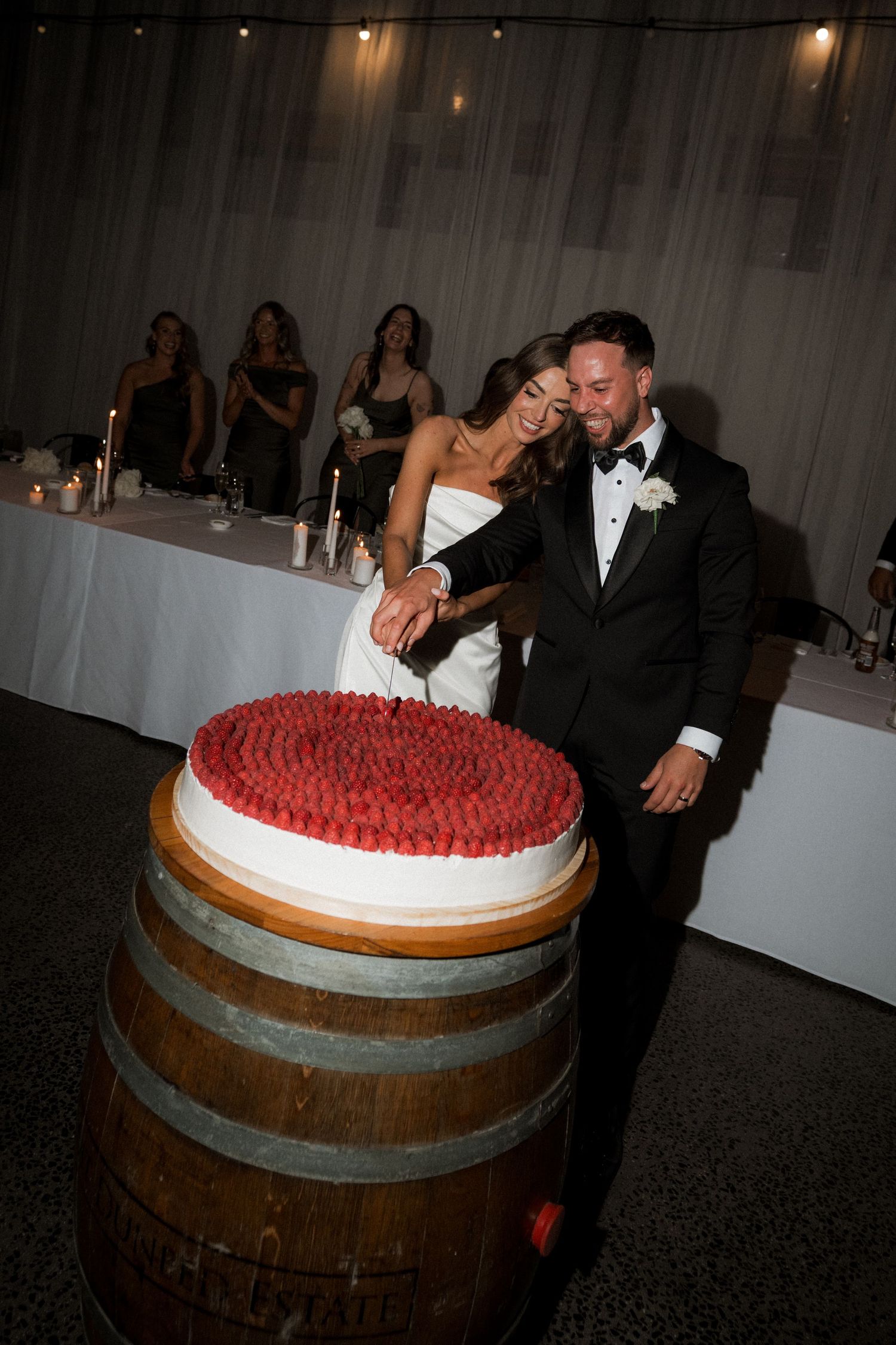 A wedding cake sits atop a rustic wooden wine barrel during an indoor reception celebration.