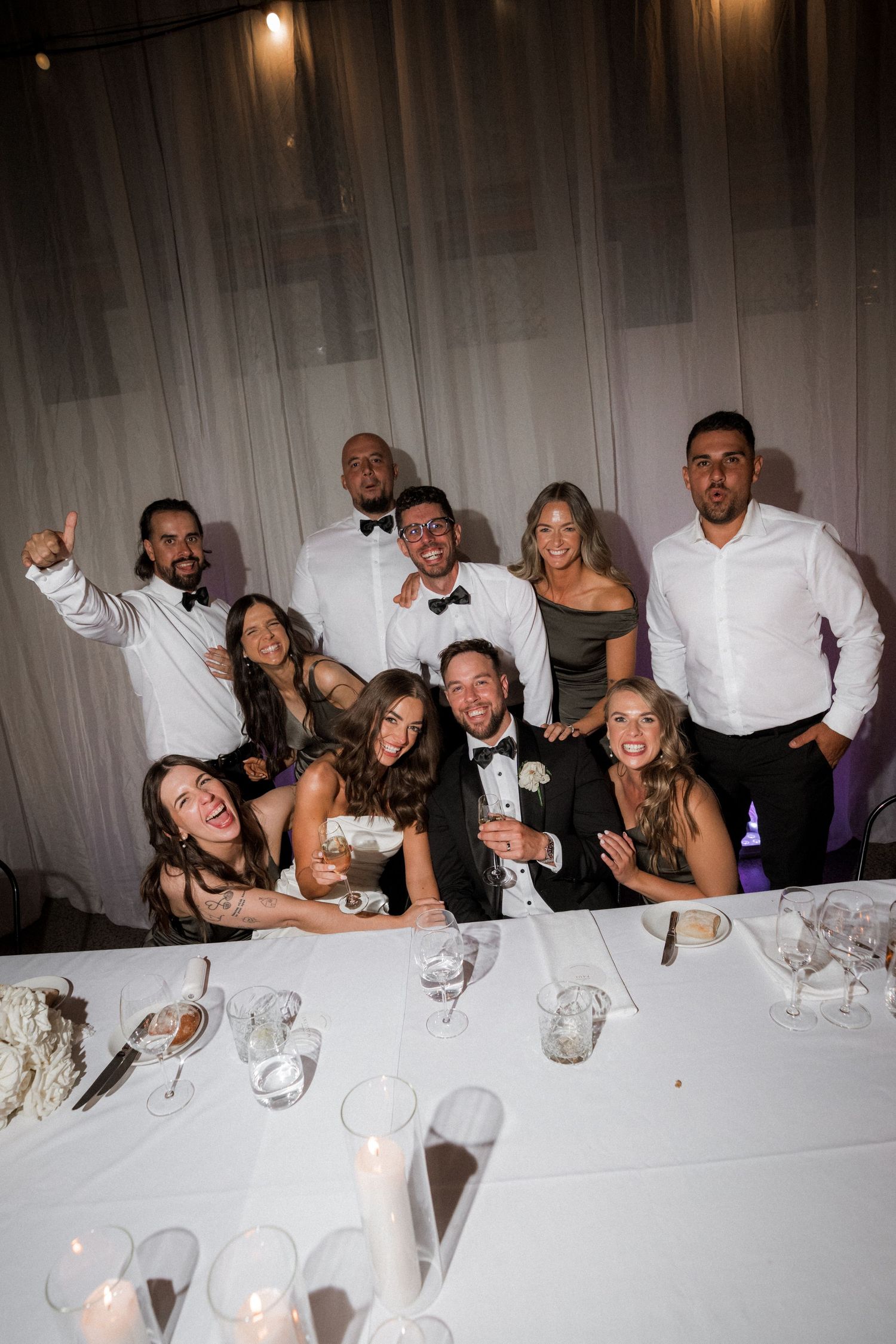 Wedding party guests pose playfully at a formal dinner table with white linens and glassware.
