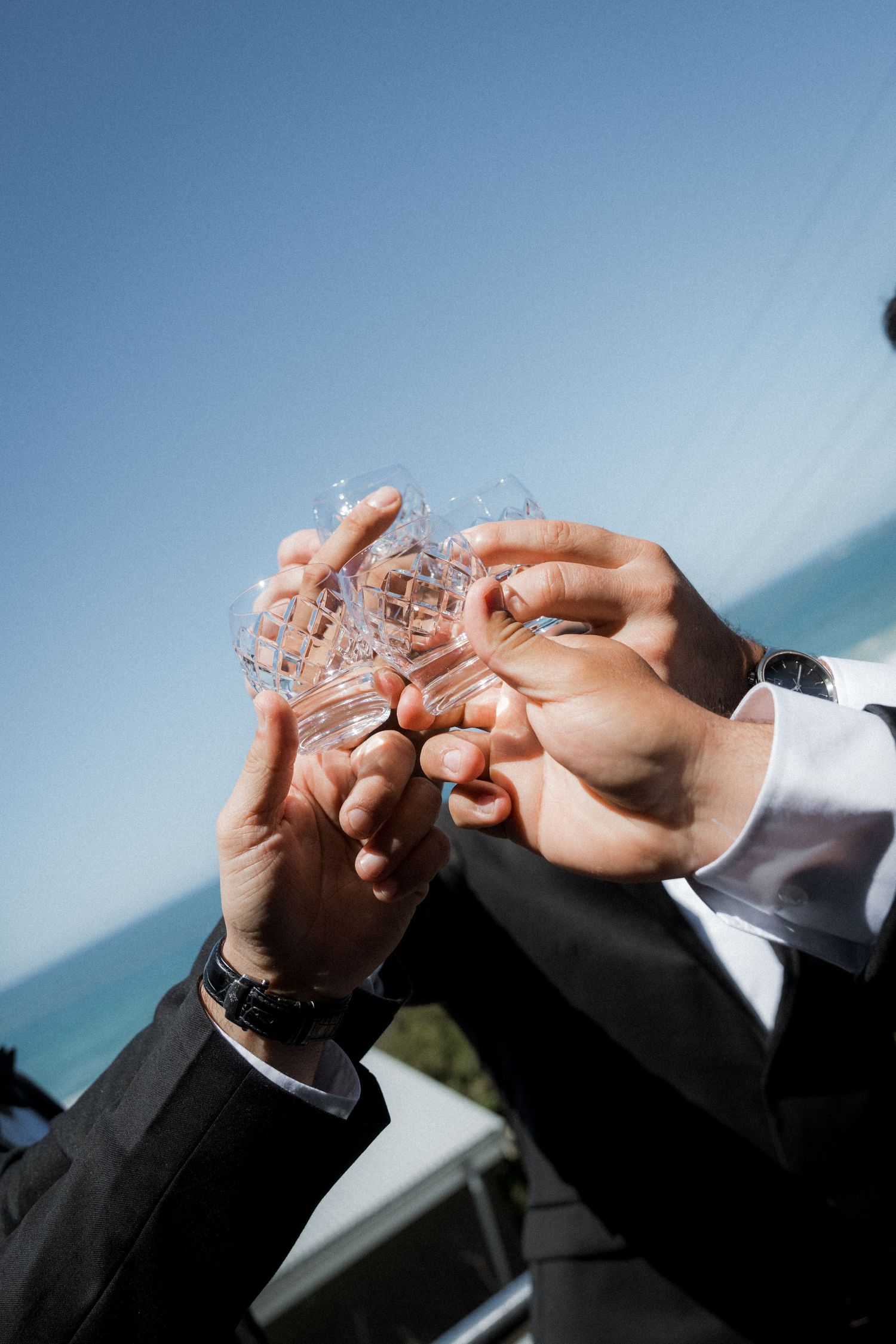 Two hands in formal attire clink crystal glasses together in a celebratory toast against a blue sky background.
