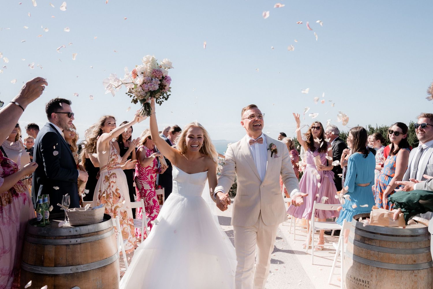 Wedding party celebrates with confetti toss against blue sky backdrop.