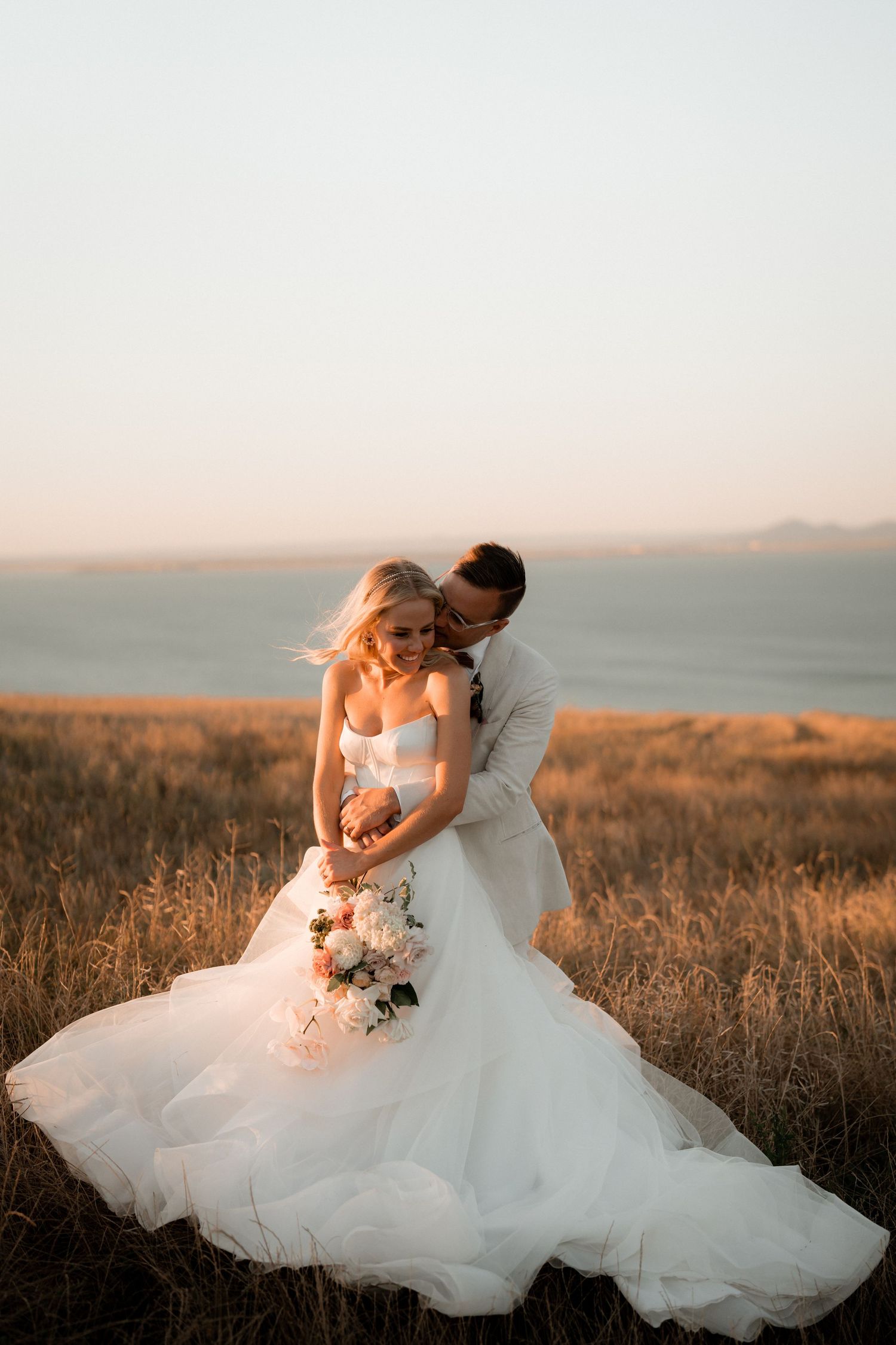 Couple embracing in flowing white wedding attire against golden sunset landscape.