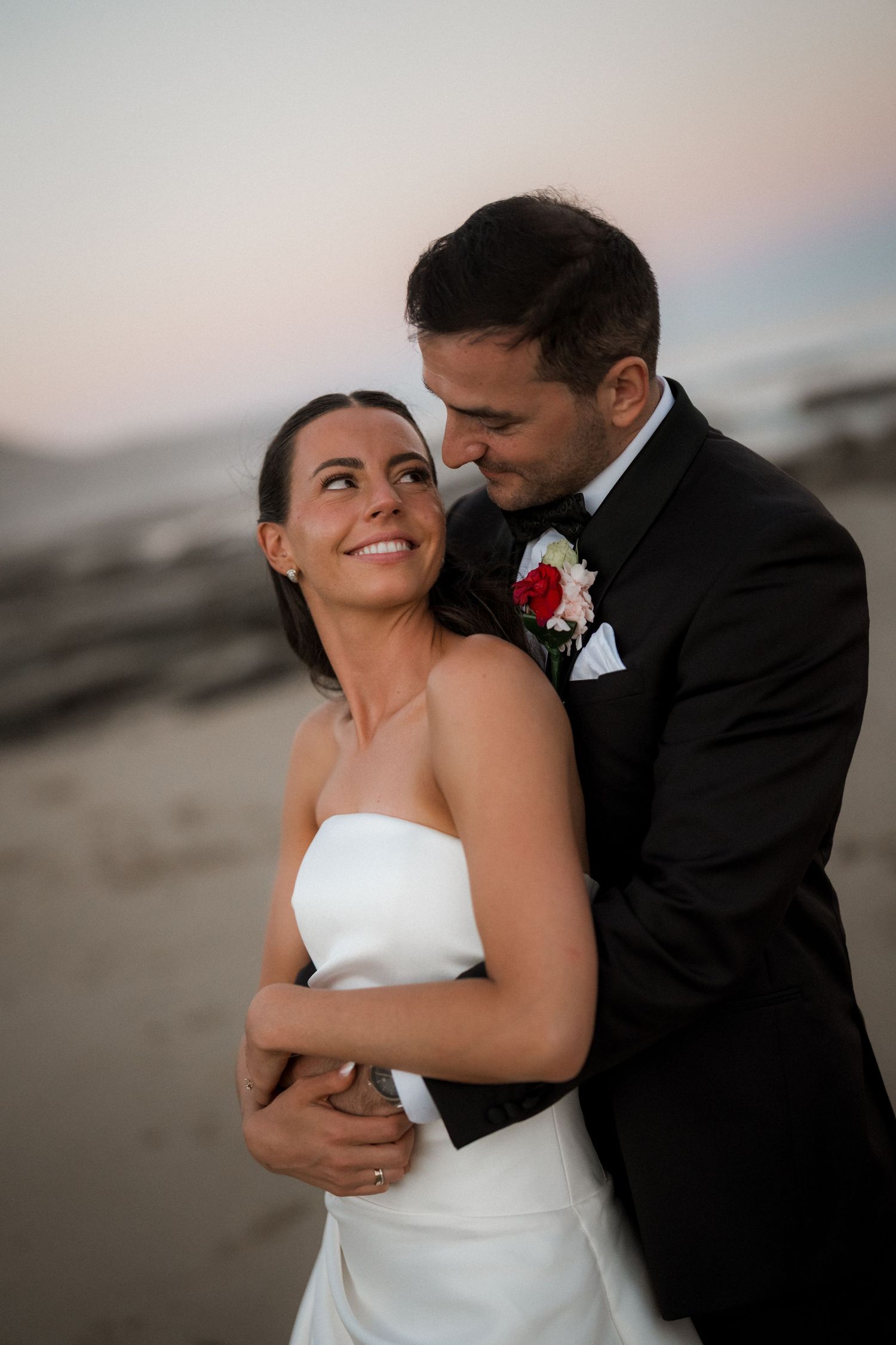 Romantic couple embrace on beach at dusk in formal wedding attire.