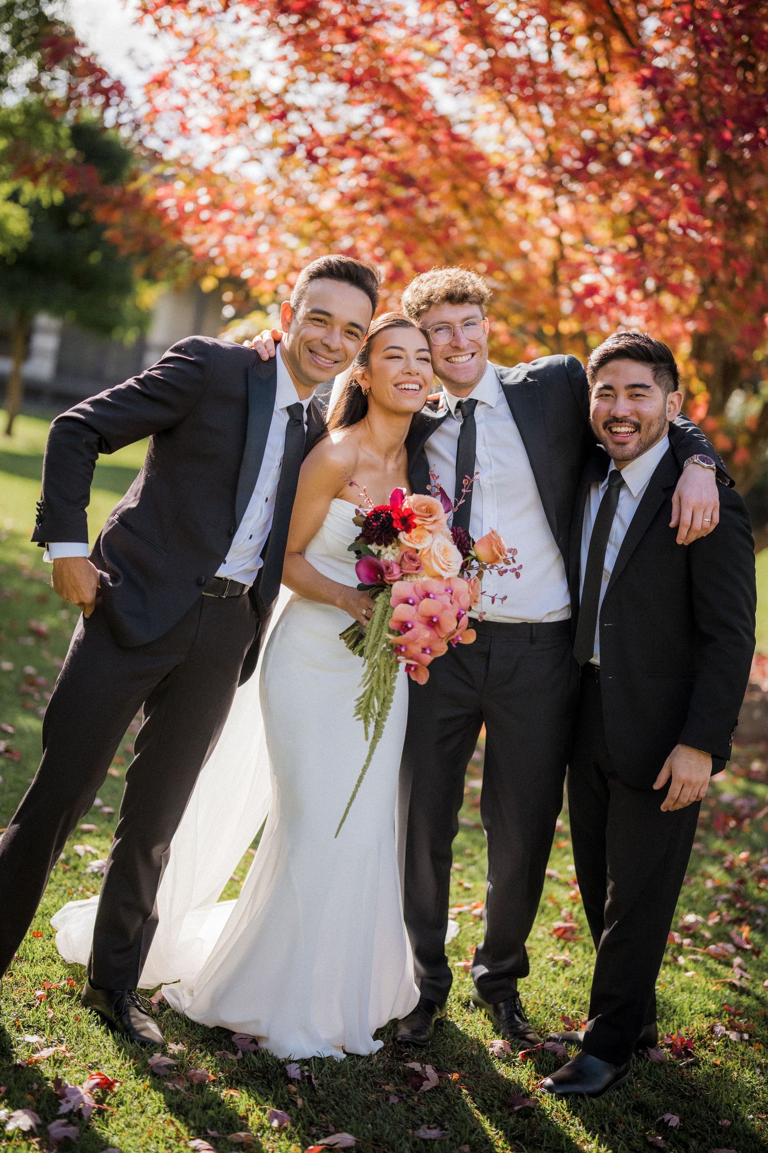 Bride in white gown holds colorful bouquet posing with three groomsmen in black suits under red autumn trees.