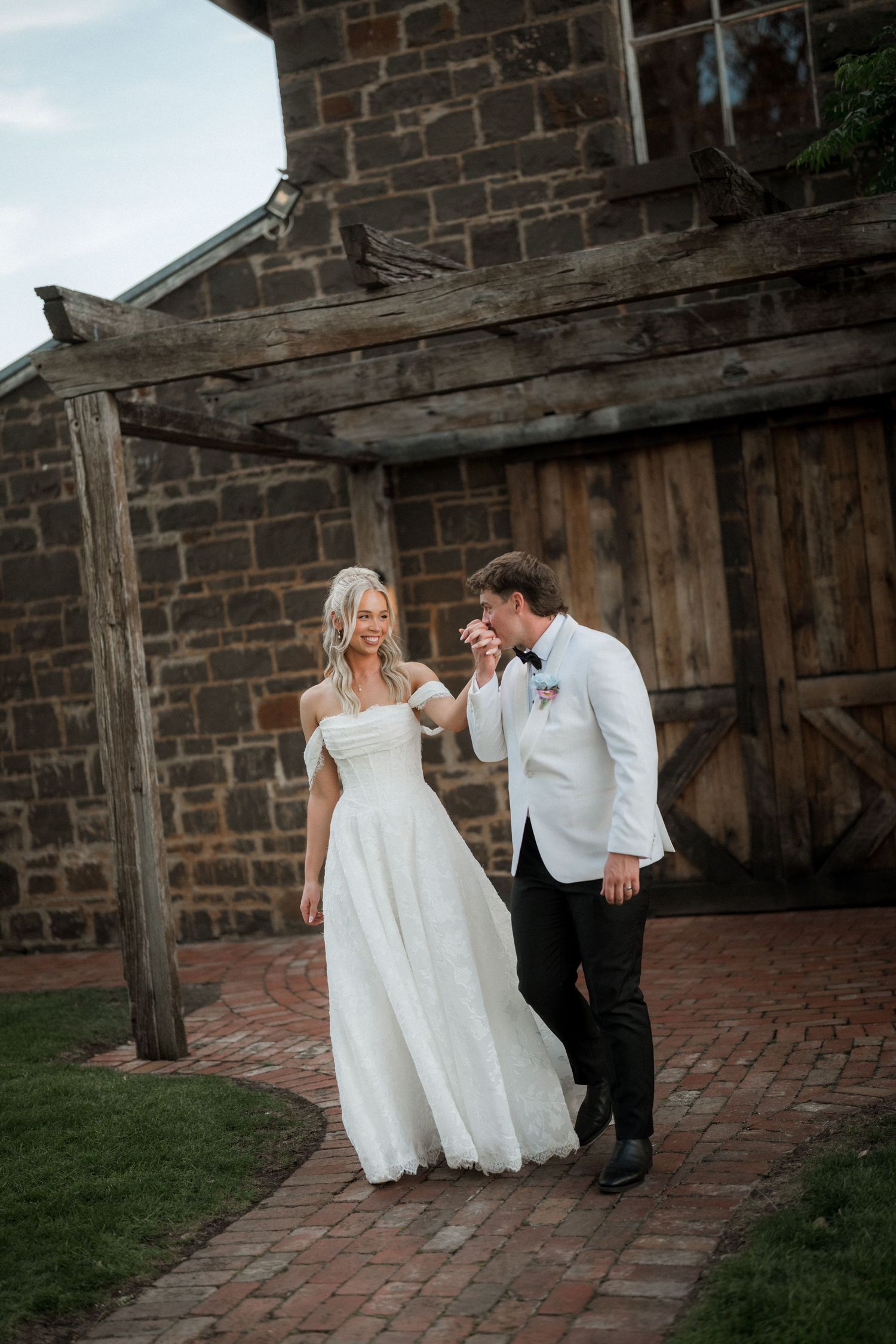 Groom in white jacket kisses bride's hand as they walk together outside a rustic stone barn wedding venue.