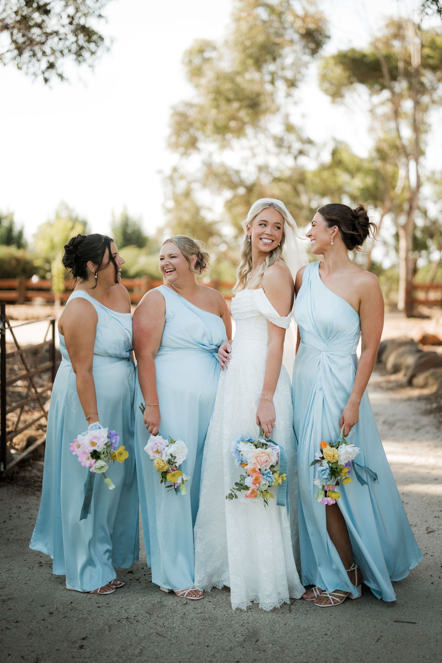 Bride with three bridesmaids in light blue dresses holding colorful bouquets outdoors.