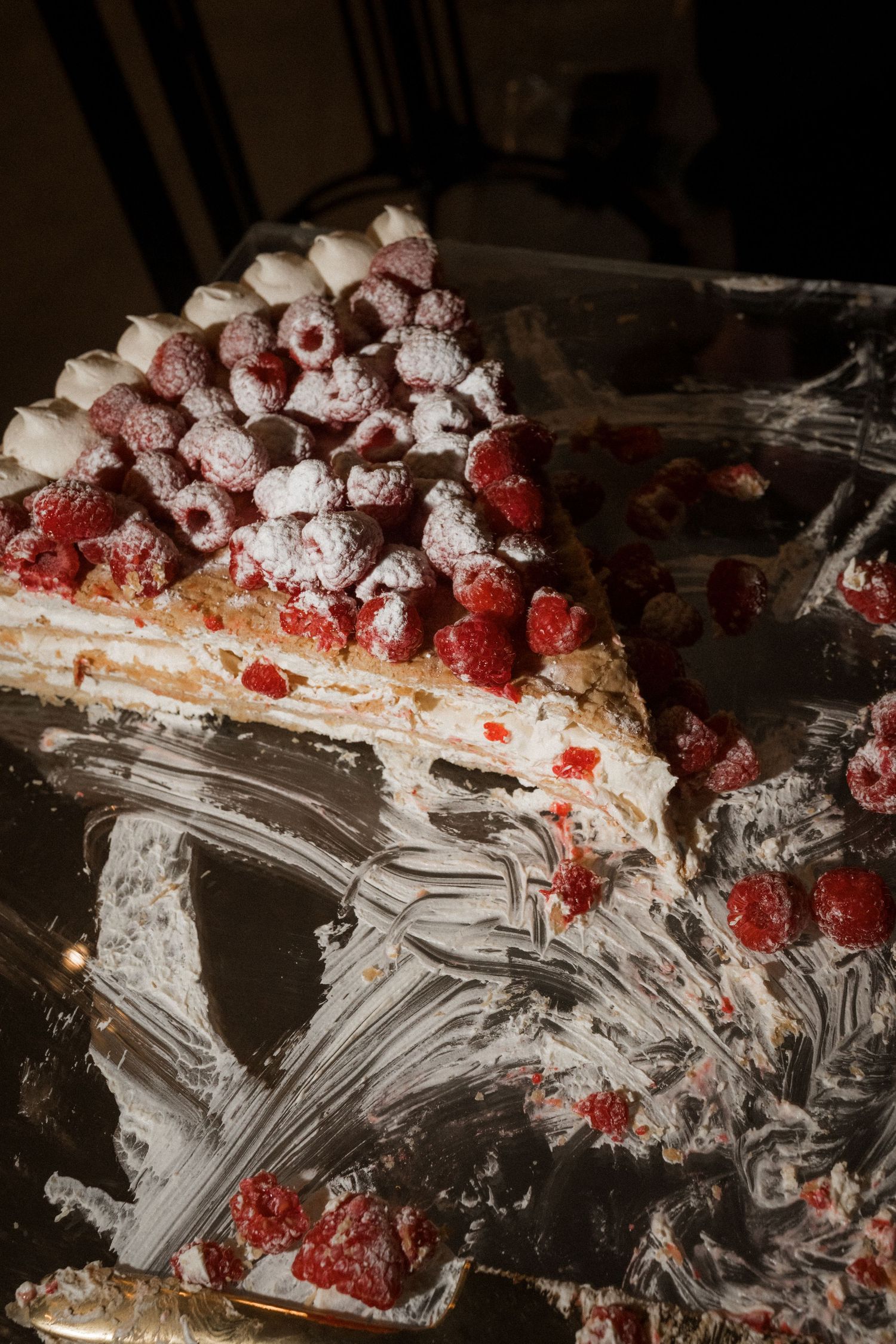 Wedding cake topped with fresh raspberries and white chocolate pieces sits on a reflective tray with cream smears and scattered berries.