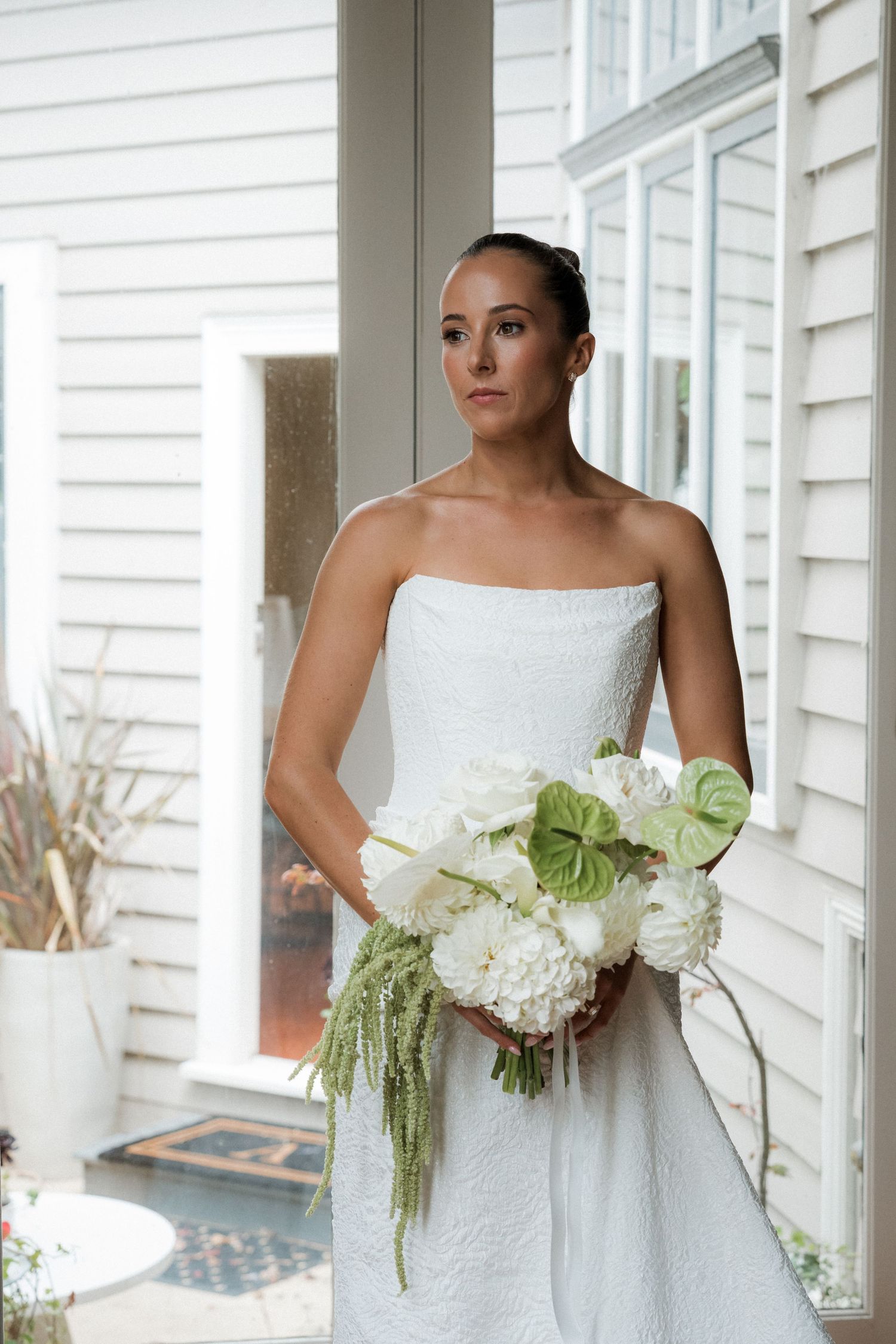 Bride in strapless white gown holding elegant white bouquet with green anthurium on a porch.